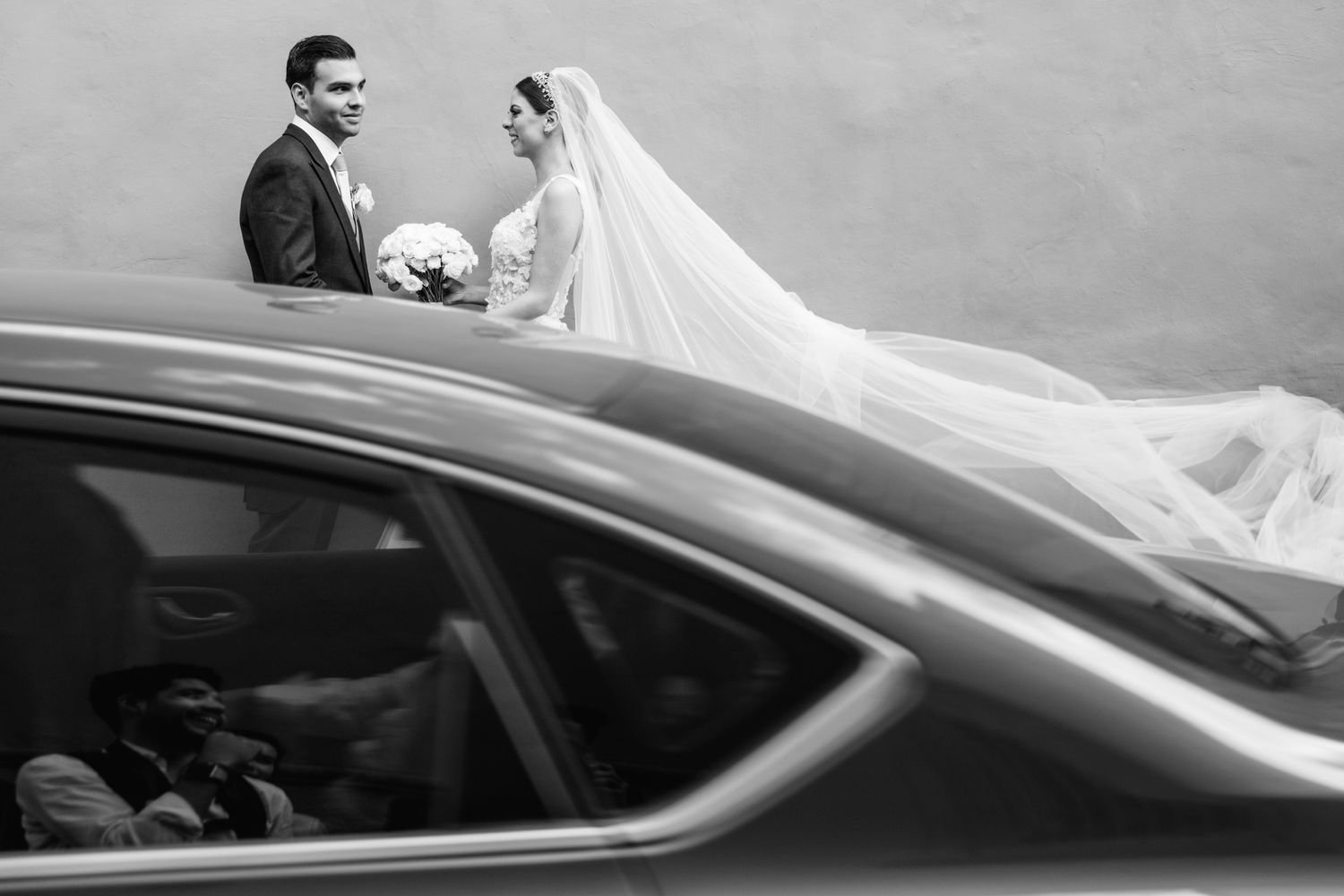 An elegant wedding couple poses beside a car with flowing veil in black and white.