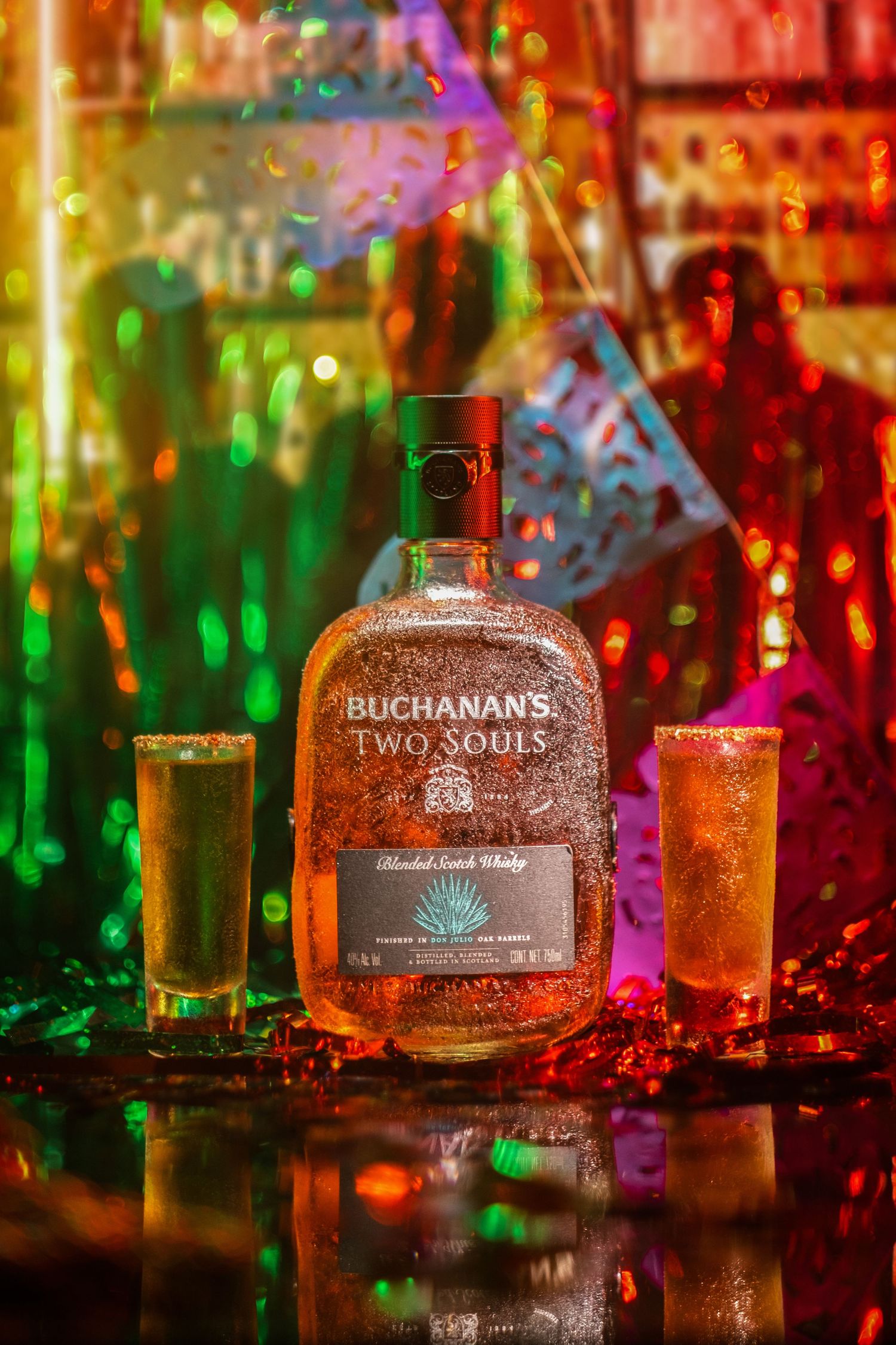 Buchanan's Two Souls whiskey bottle displayed on a bar with colorful bokeh lights creating a festive nightclub atmosphere.