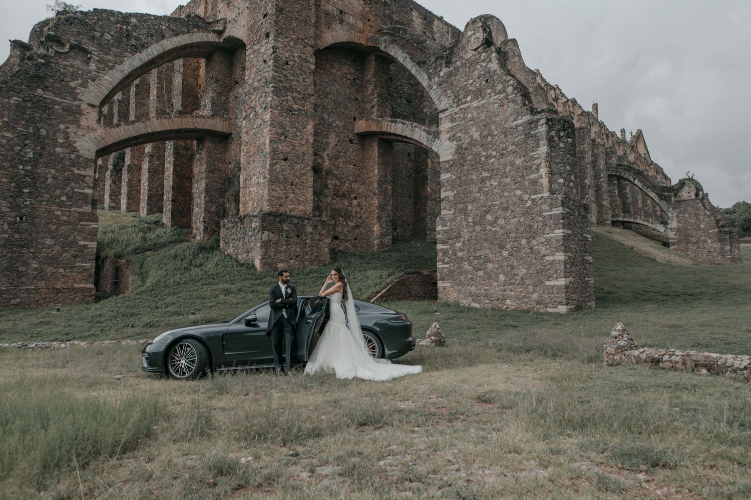 Newlyweds pose with vintage car in front of dramatic stone ruins of historic abbey on overcast day.