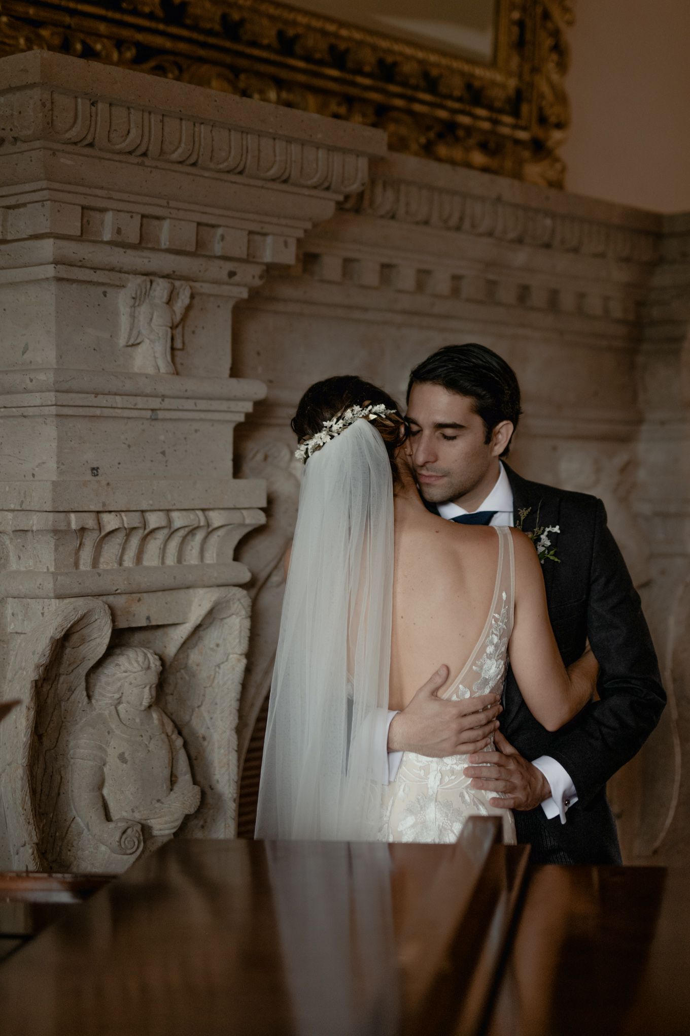 A wedding couple embraces intimately in front of an ornate stone wall with architectural details and carved reliefs.