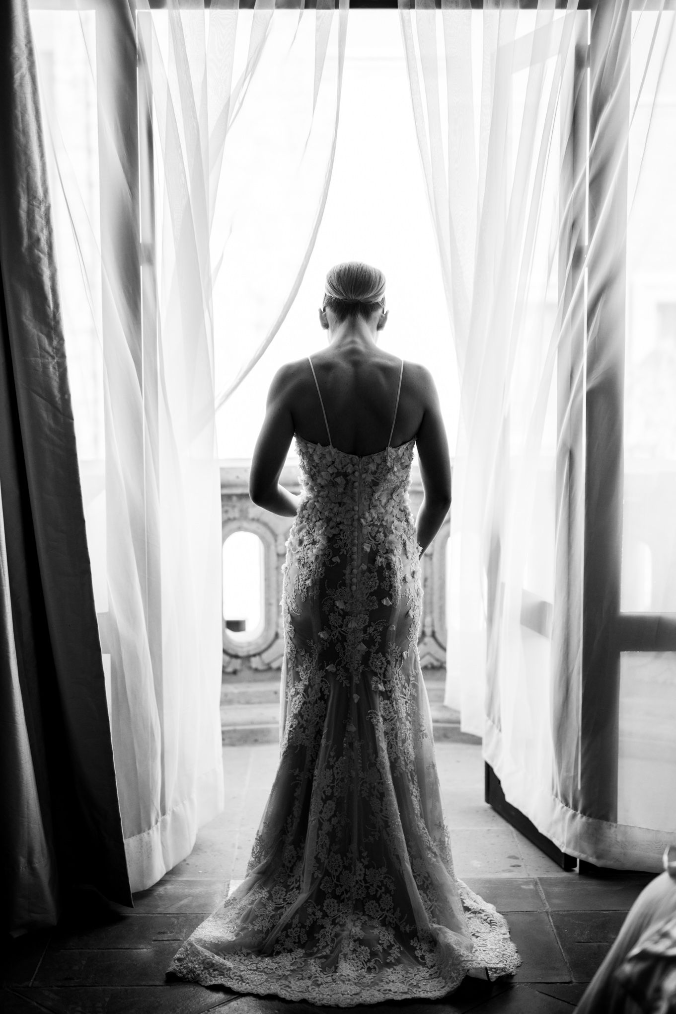 Black and white photo of a bride in lace wedding gown silhouetted against sheer curtains in dramatic lighting.