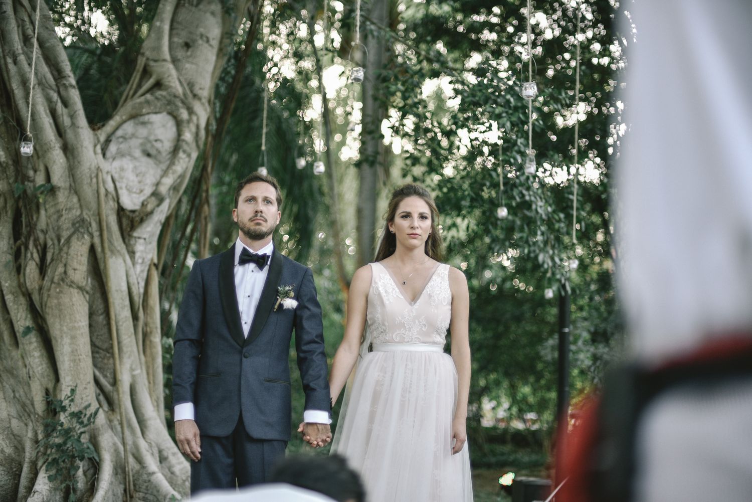 A bride and groom hold hands during their romantic outdoor wedding ceremony beneath tall trees in a garden setting.