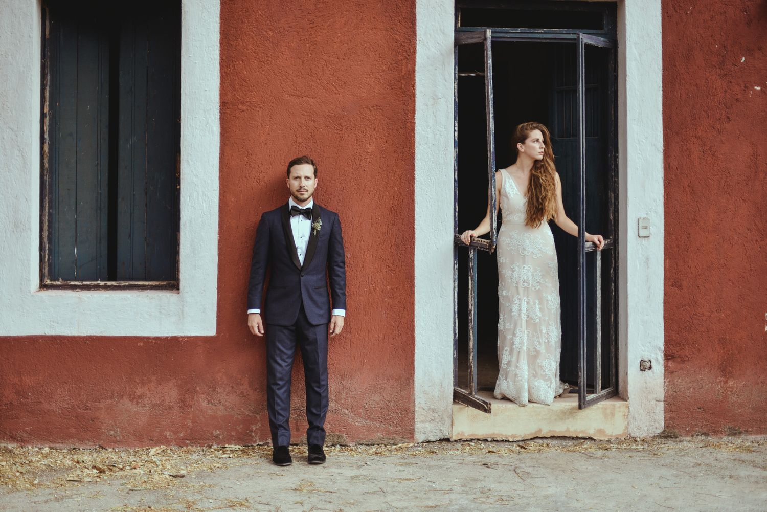 A couple in formal wedding attire poses against a rustic red wall outside an old Italian villa doorway.