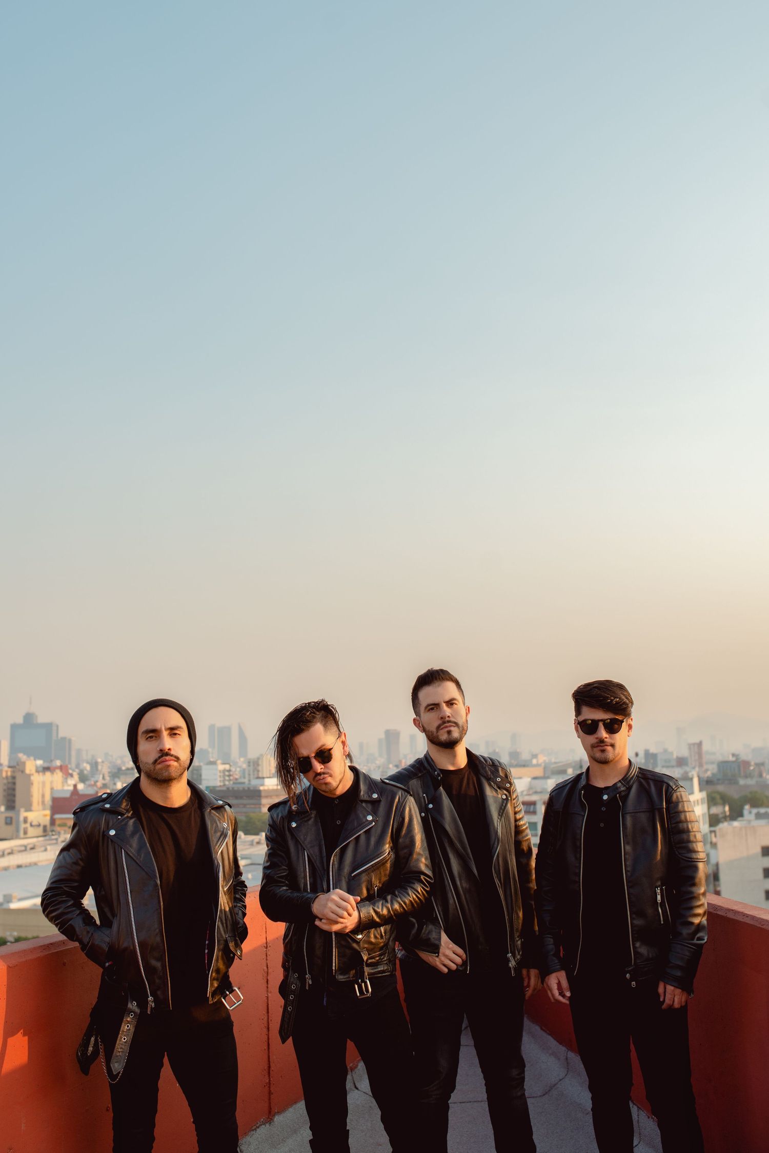 Rock band in leather jackets poses on rooftop against city skyline at sunset.