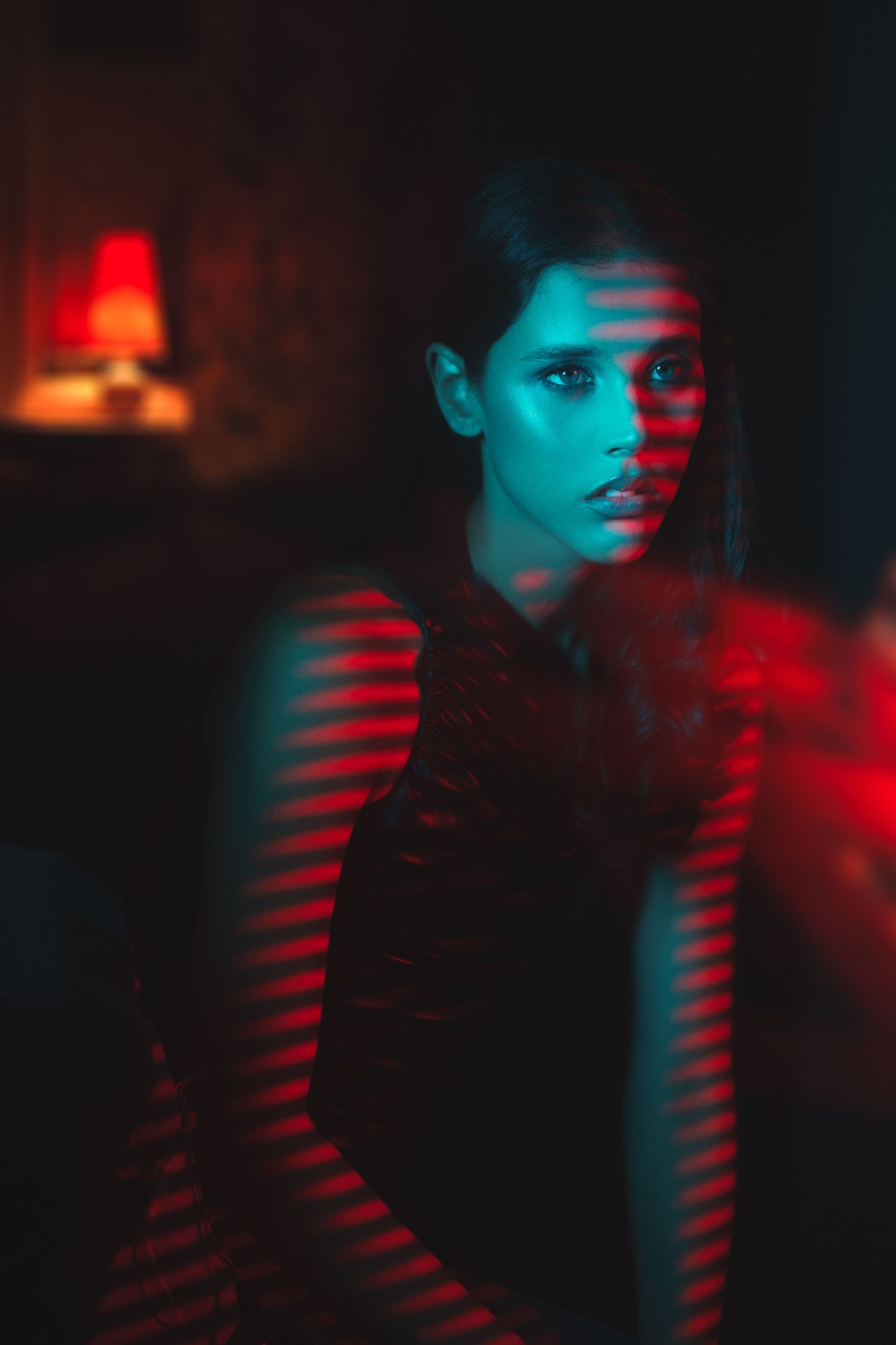 Dramatic portrait with red and blue lighting casting striped shadows in dark moody atmosphere.