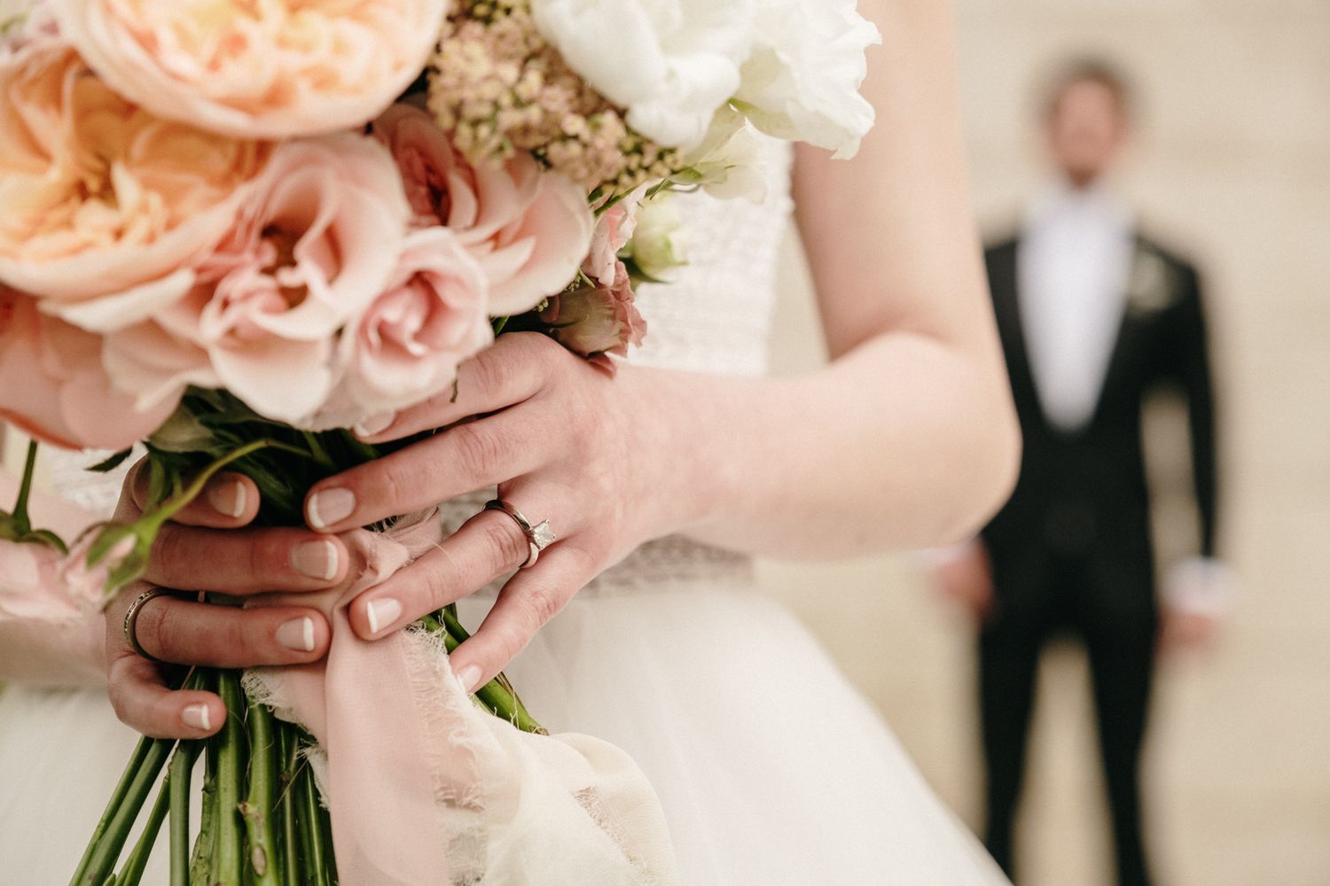 Close-up shot of wedding rings and bouquet with couple in soft focus background.