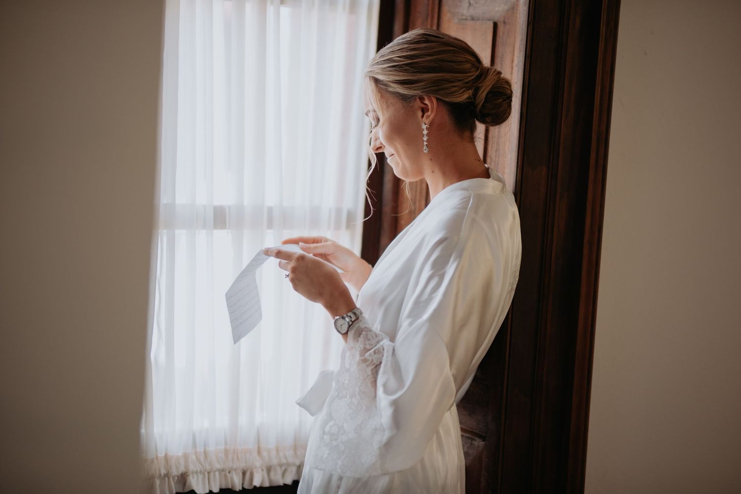 Figure in white robe reads letter by window during wedding morning preparations.
