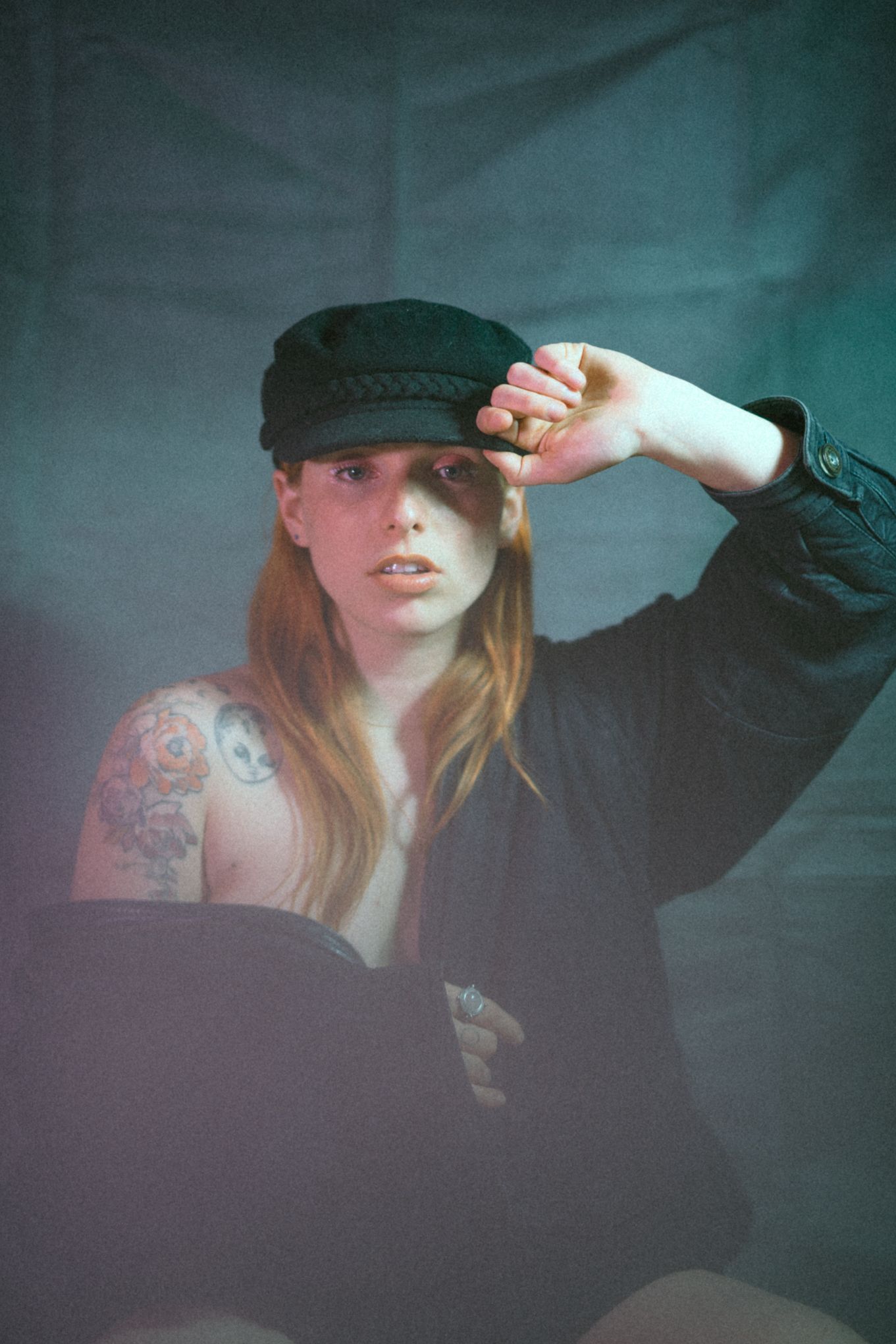 Moody portrait with newsboy cap and visible tattoo against teal background.