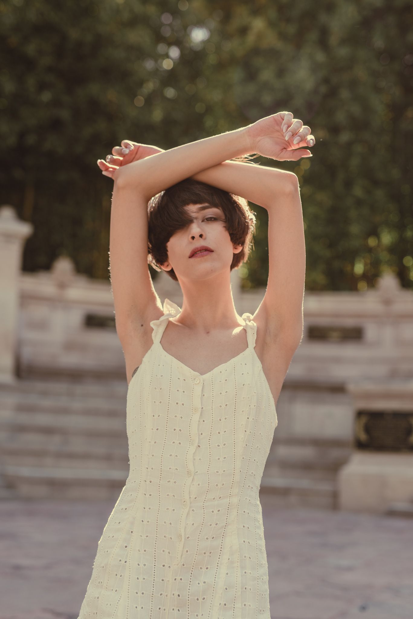 Woman in a white eyelet sundress poses gracefully with raised arms against an outdoor backdrop.