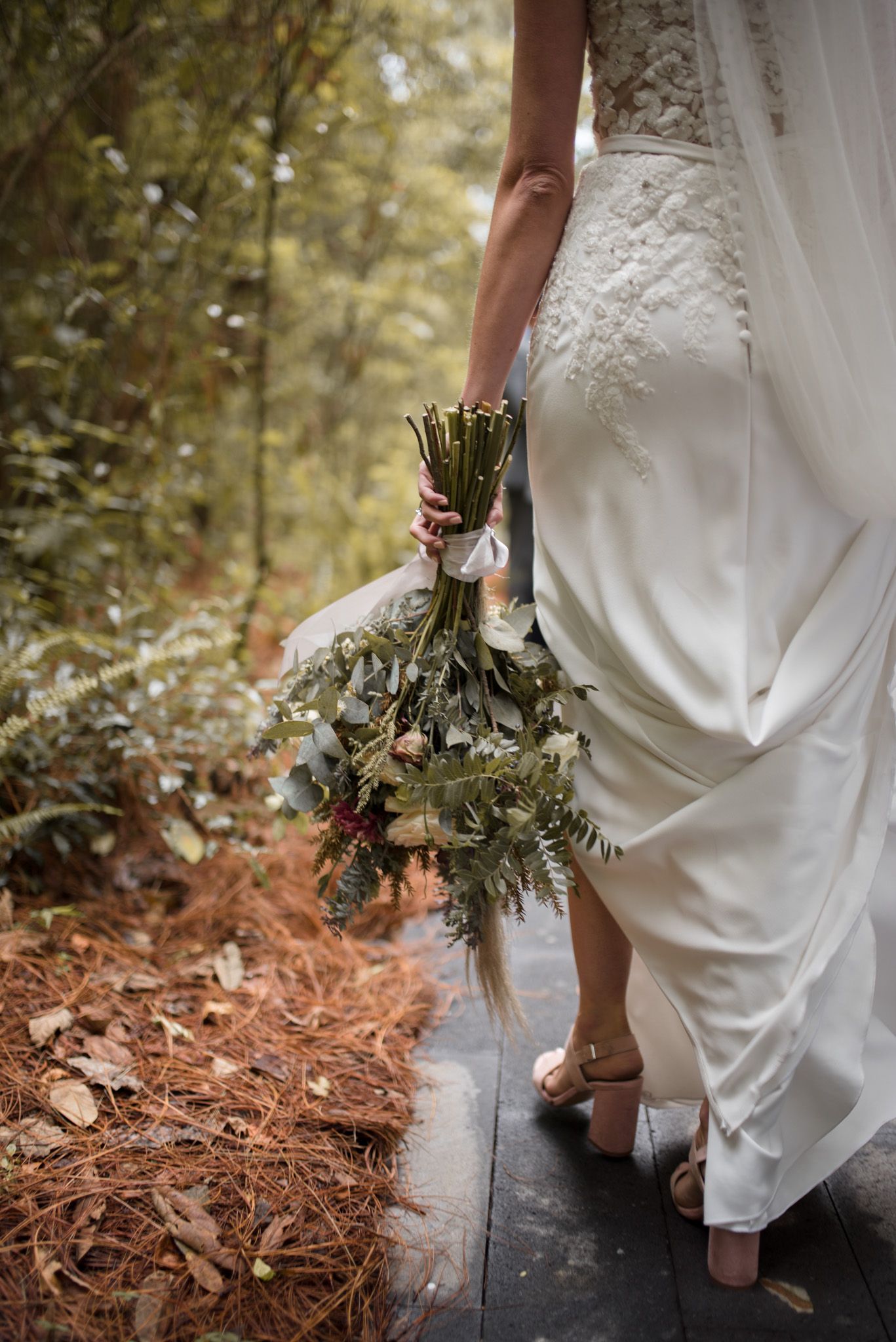 Back view of bride in flowing white lace dress holding rustic bouquet on wooden path through autumn woods.