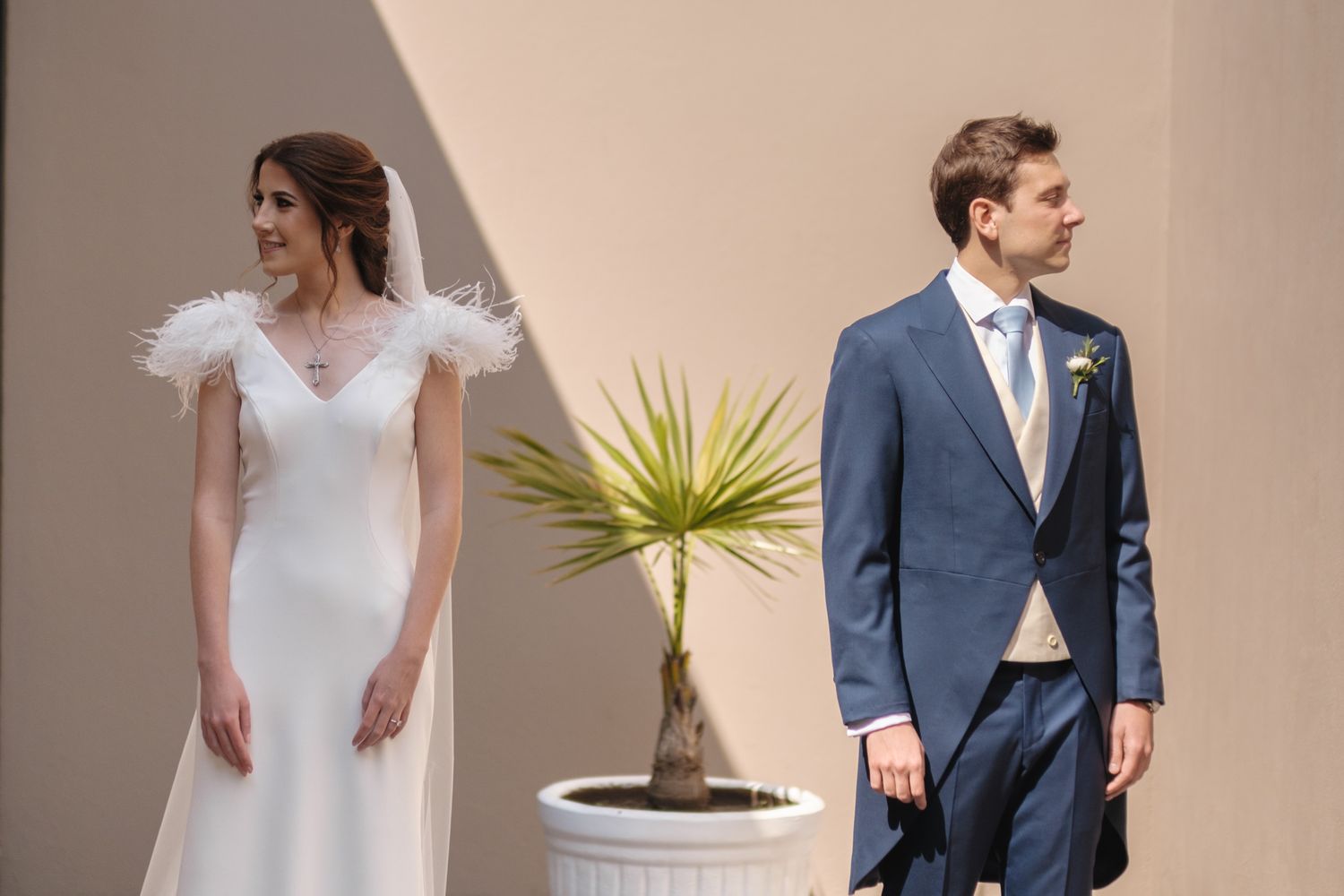 A couple in wedding attire stand apart near a potted plant against a beige wall on their special day.