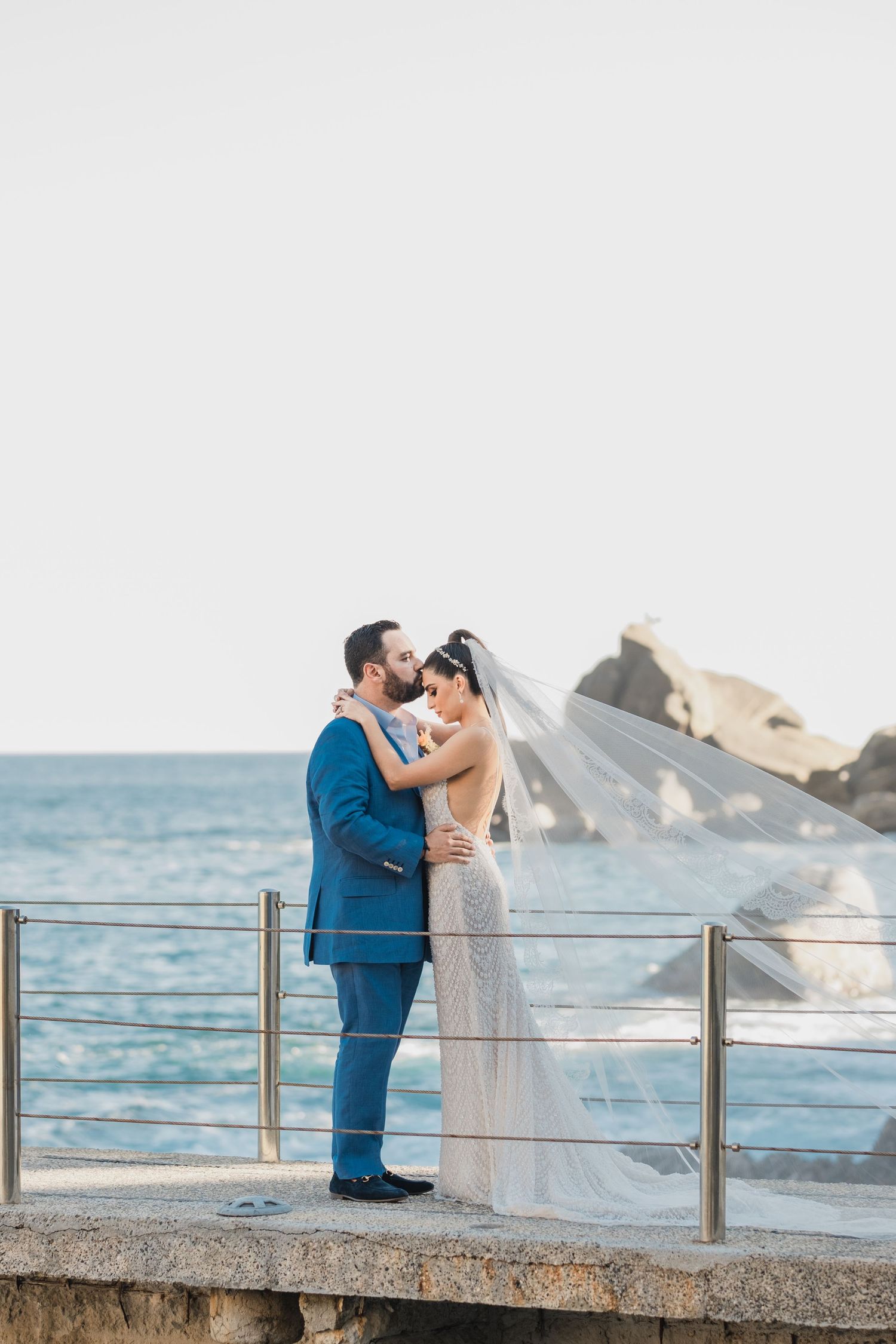 A couple shares a romantic seaside moment on a stone pier as the bride's veil flows in the breeze.