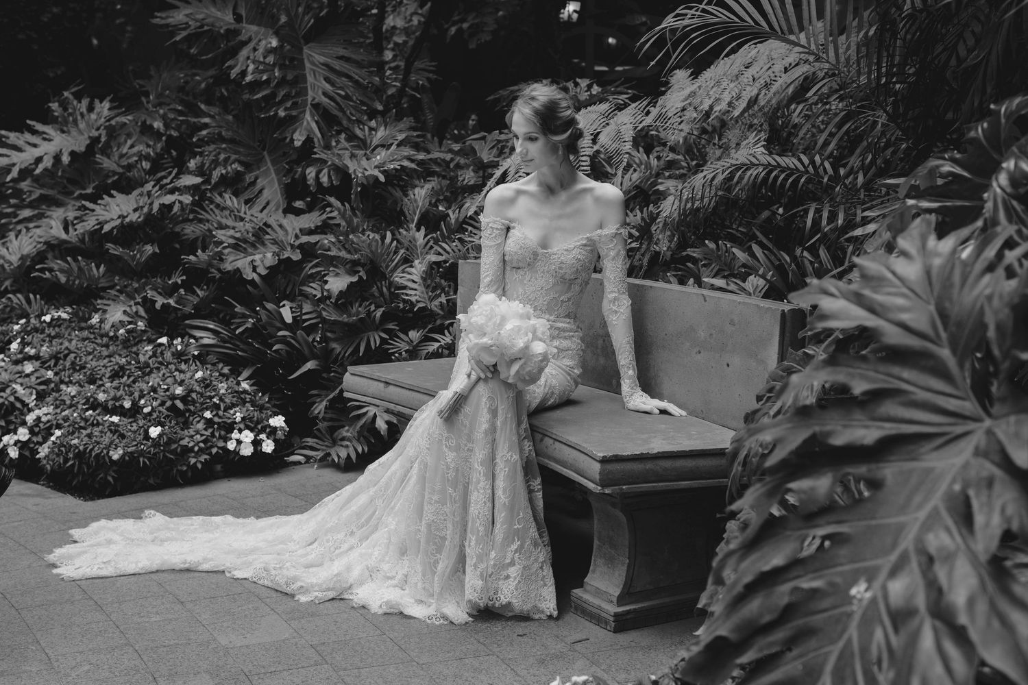 Artistic black and white photo of a bride in lace sitting on a garden bench surrounded by tropical plants.