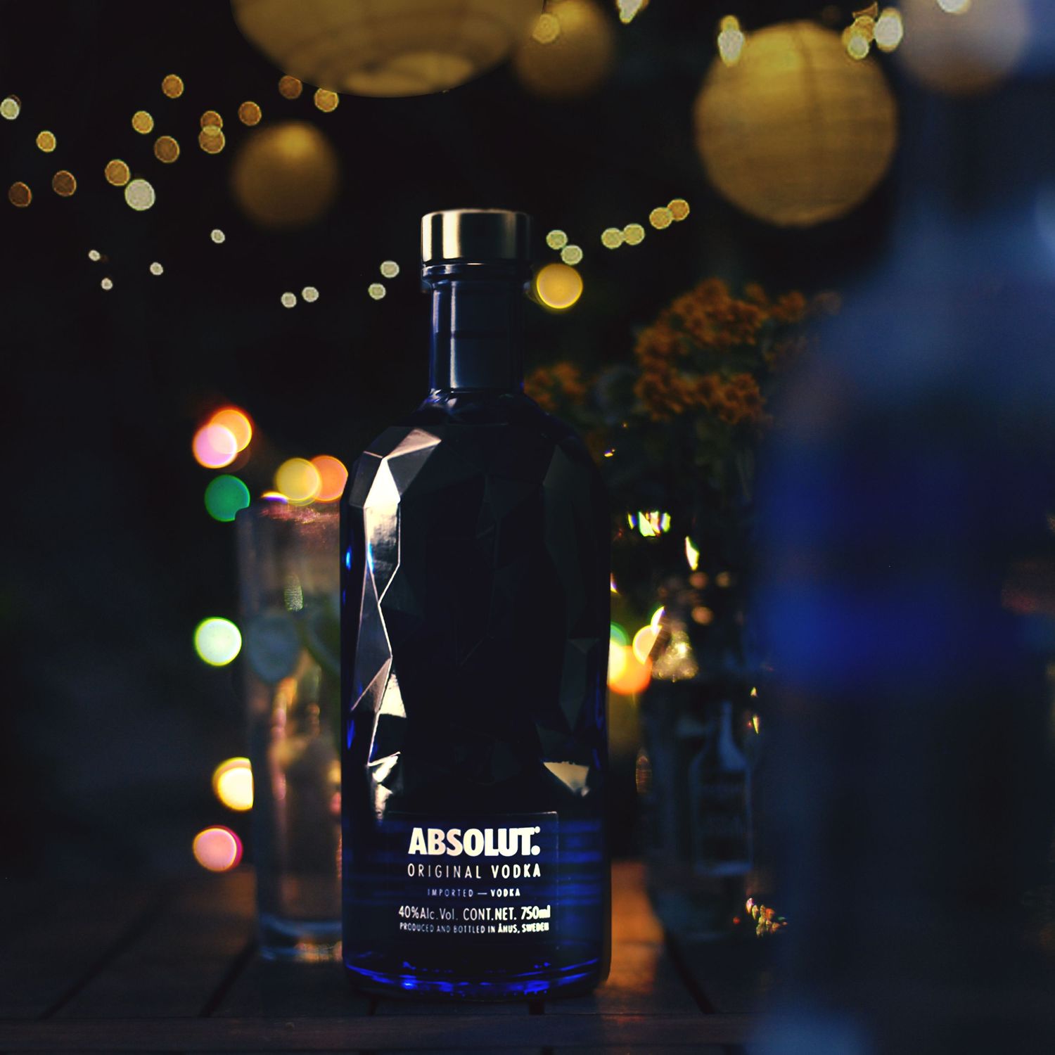 Blurred bokeh lights create a festive atmosphere around dark bottles of Absolut vodka in moody nighttime setting.