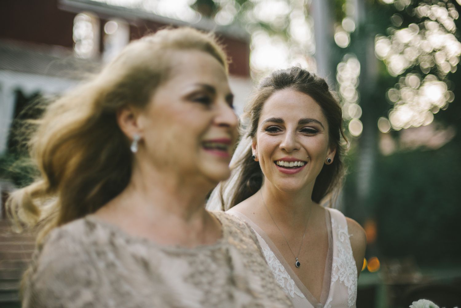 Two people sharing laughter and joy at an outdoor event, wearing light colored elegant dresses in natural lighting.