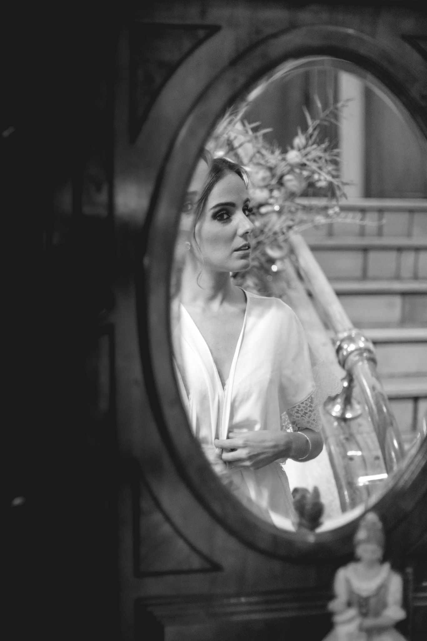 Black and white reflection in oval mirror shows elegant figure in white dress holding bouquet during bridal preparation.