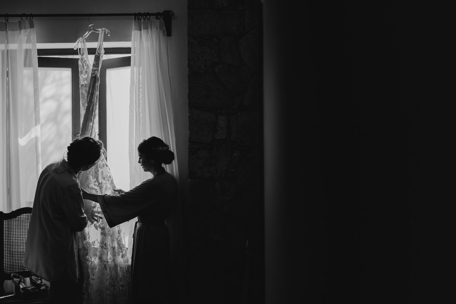 A black and white silhouette photo of two figures standing by a bright window with sheer curtains.