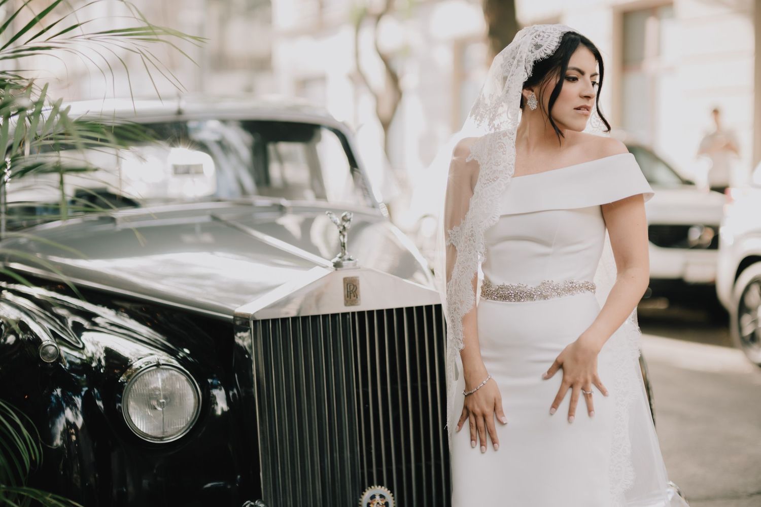 Elegant bride in off-shoulder white gown poses next to classic black Rolls Royce on city street.