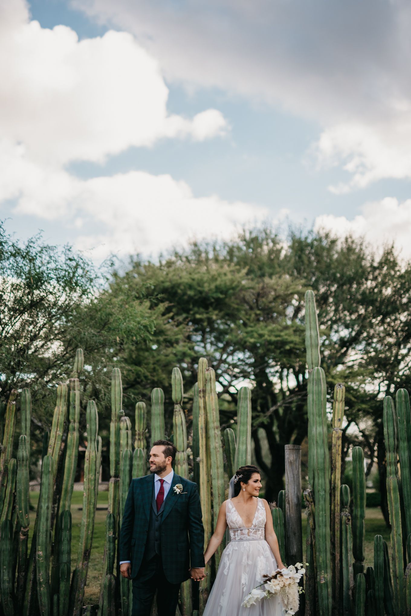 Wedding couple poses in front of tall desert cactus garden at outdoor venue under cloudy sky.