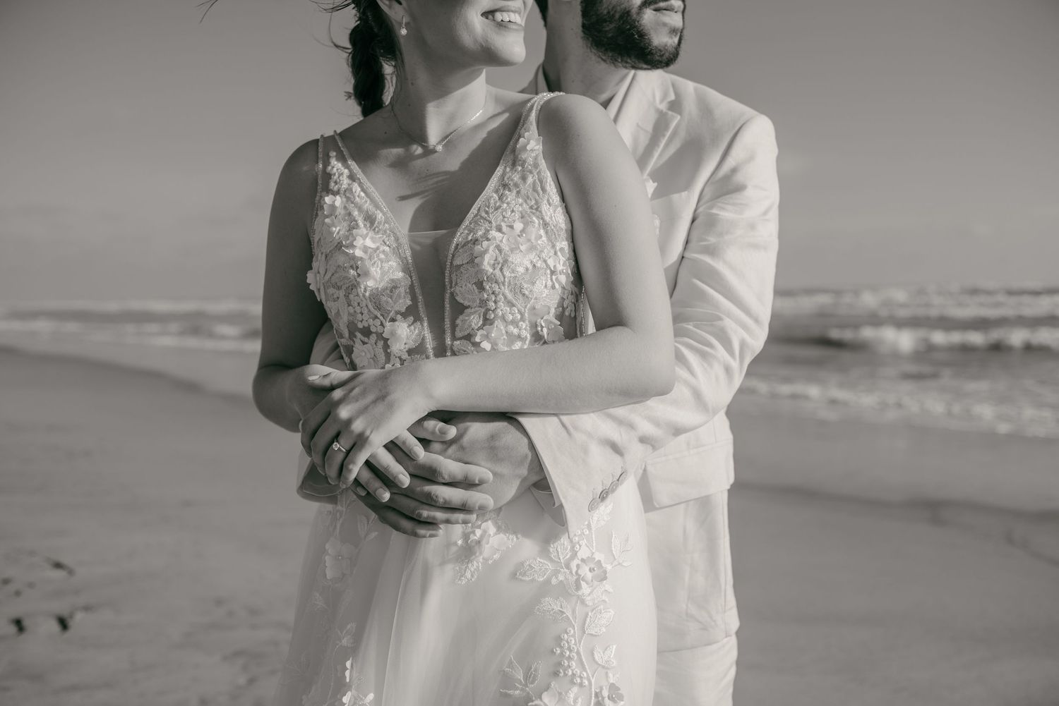 A romantic black and white photo of a couple embracing on the beach, wearing wedding attire.