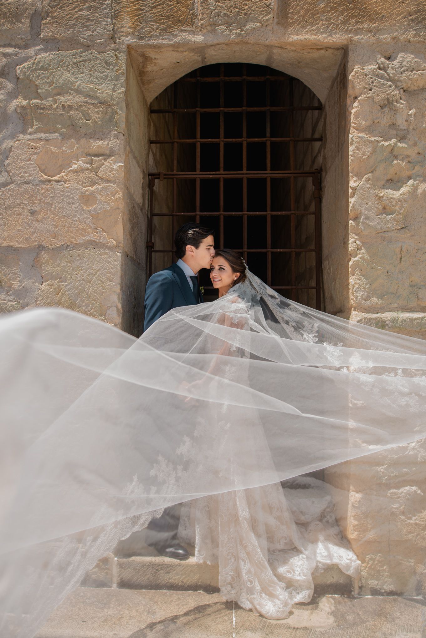 Elegant wedding couple embracing near stone archway while flowing white veil creates romantic movement in the breeze.