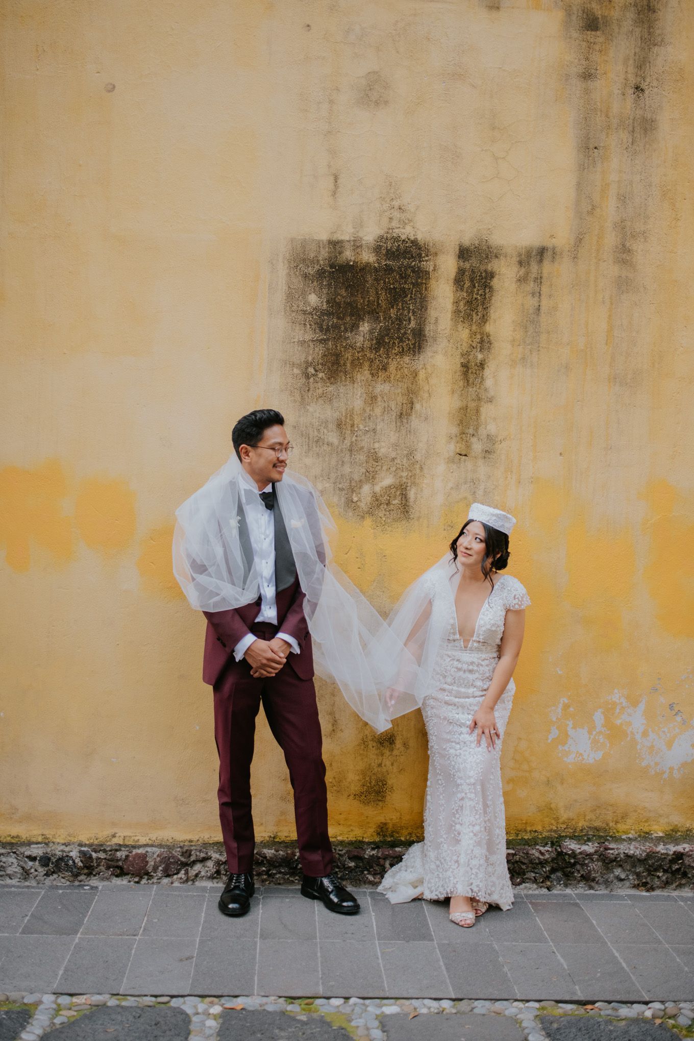 Modern wedding couple poses casually against textured yellow wall.