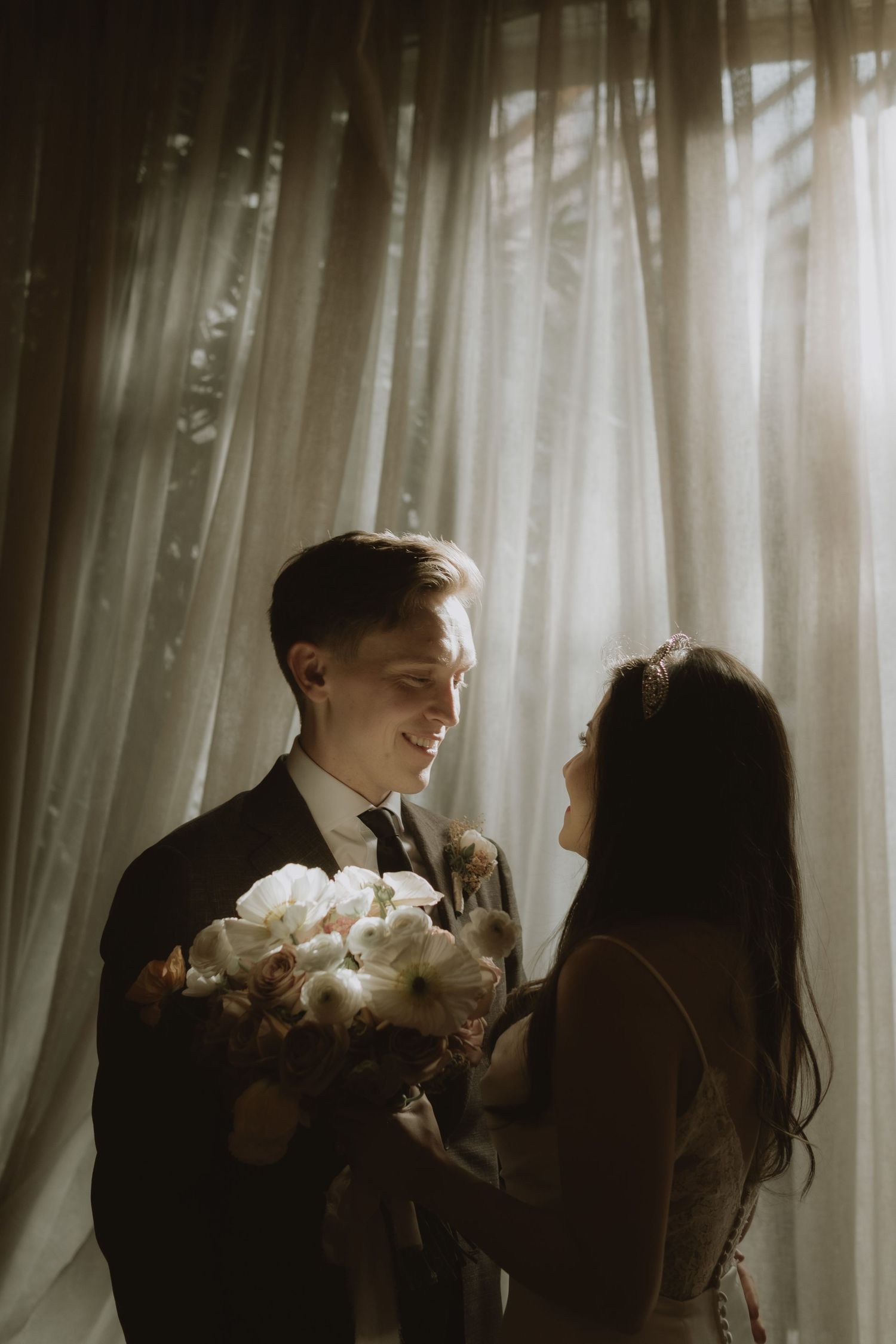 Romantic silhouette of couple sharing a moment with white flowers near sheer curtains.
