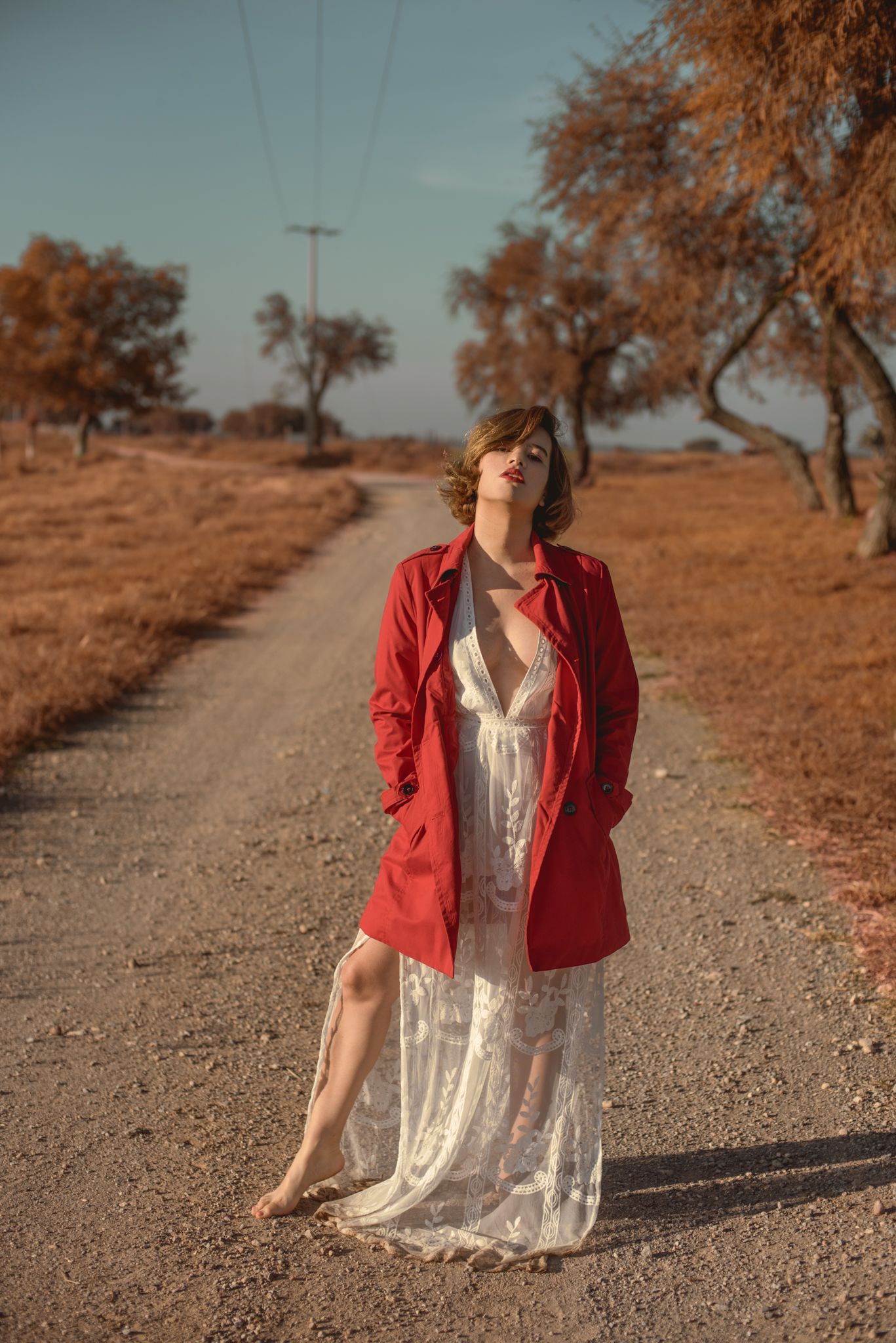 Someone in white dress and red jacket poses on dusty rural road with autumn trees.