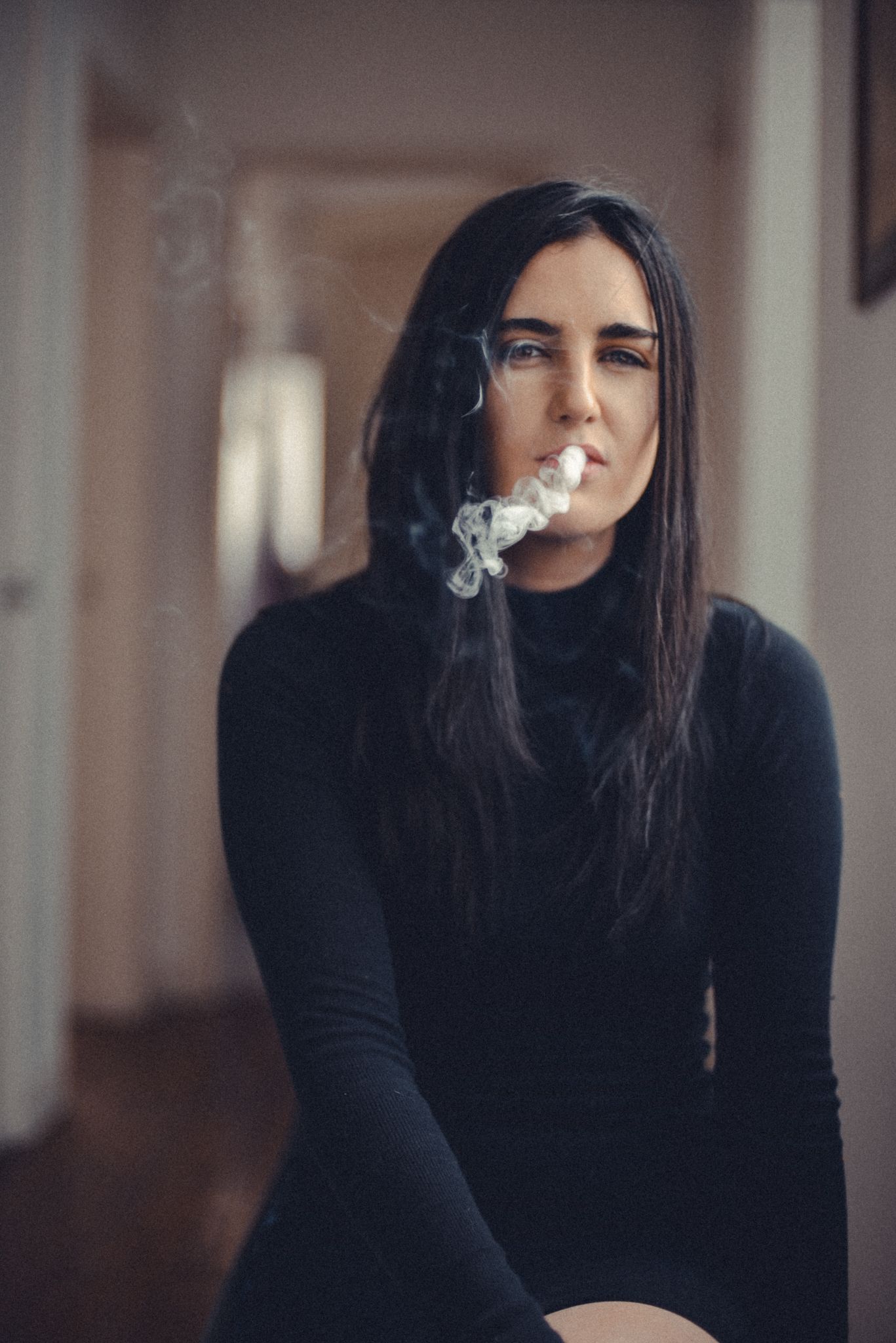 Person in black sweater exhaling smoke in moody indoor setting.