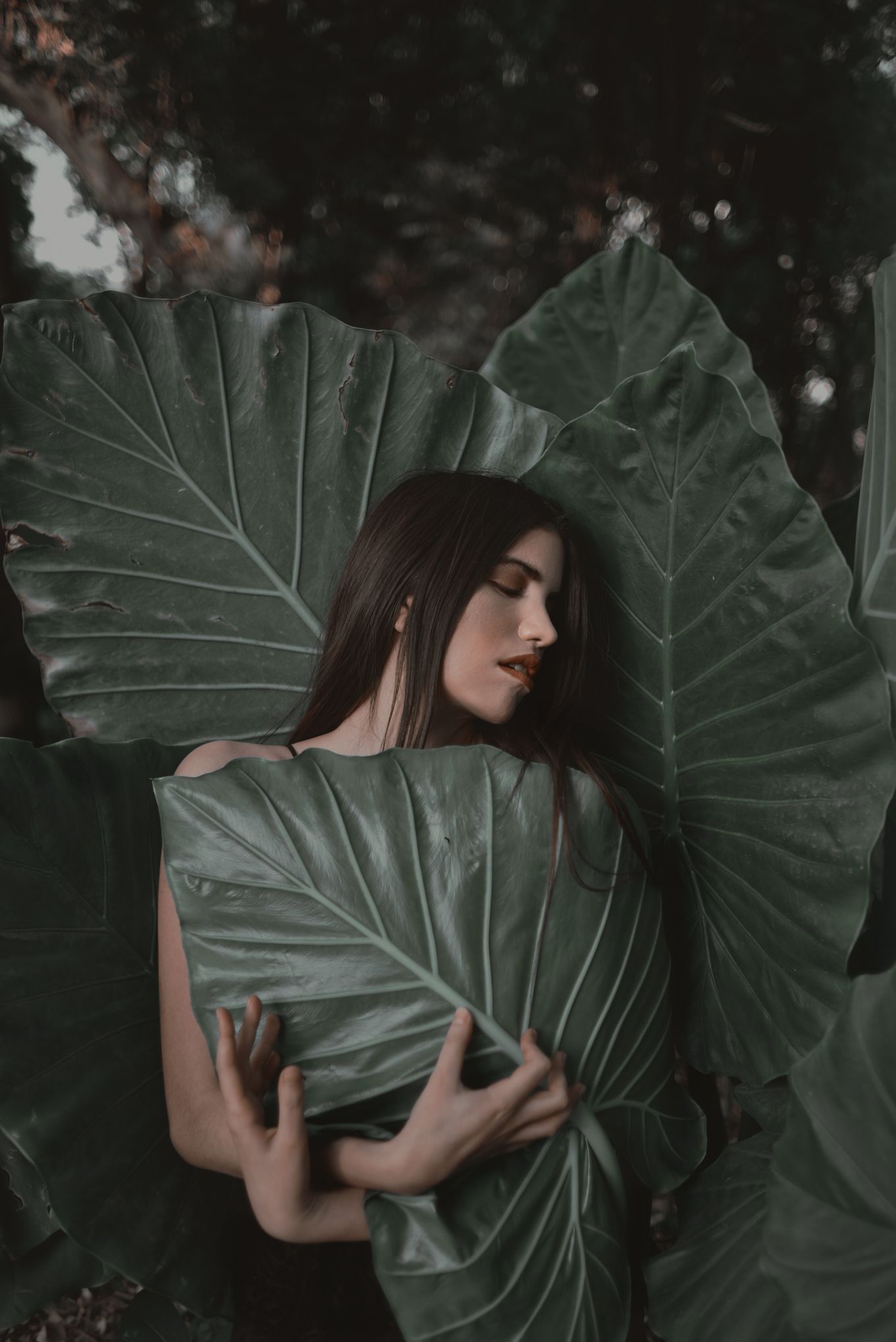 Artistic portrait of woman posing with large green tropical leaves in moody lighting.
