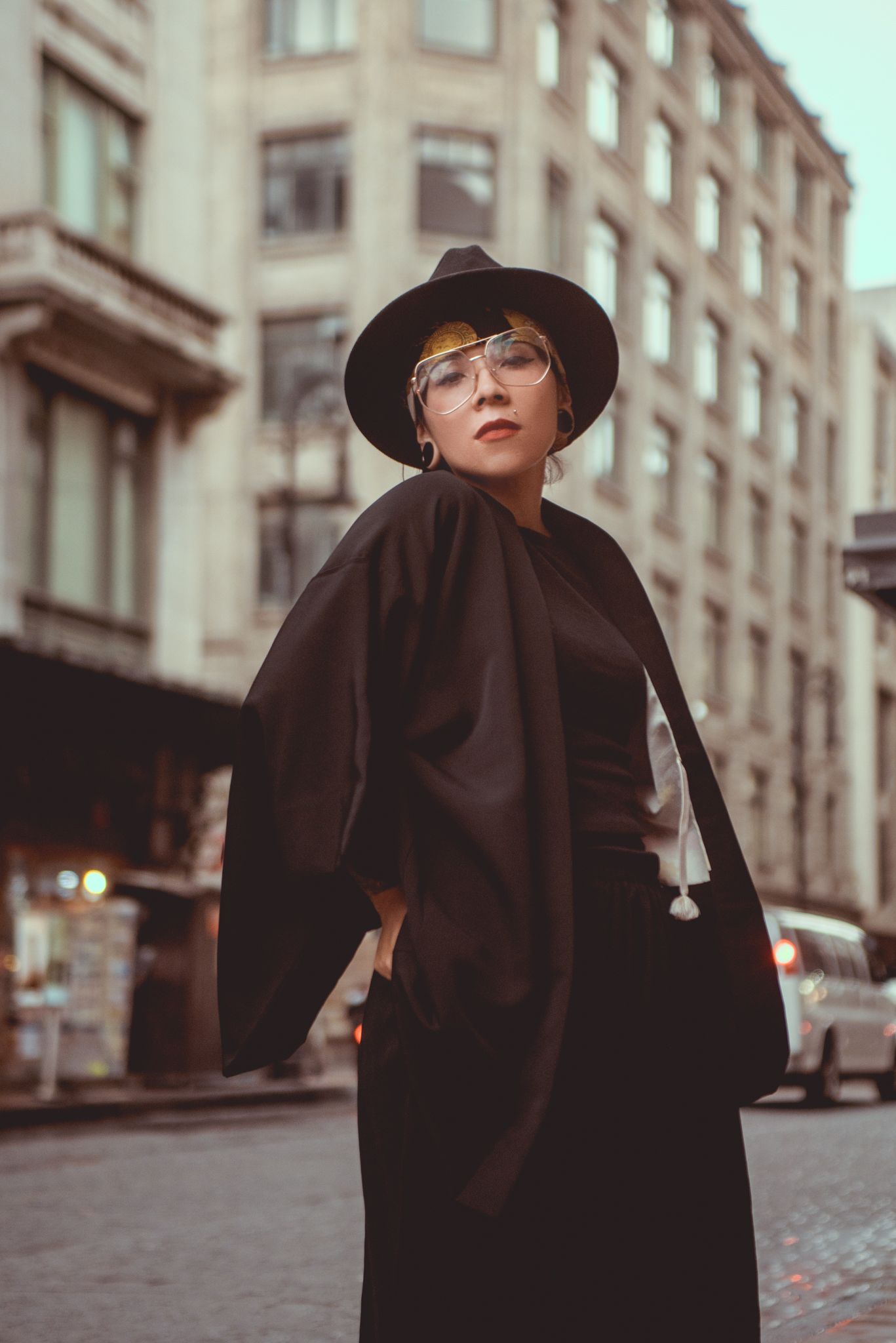 Fashion portrait of someone in vintage-style black coat and fedora hat against urban architecture.