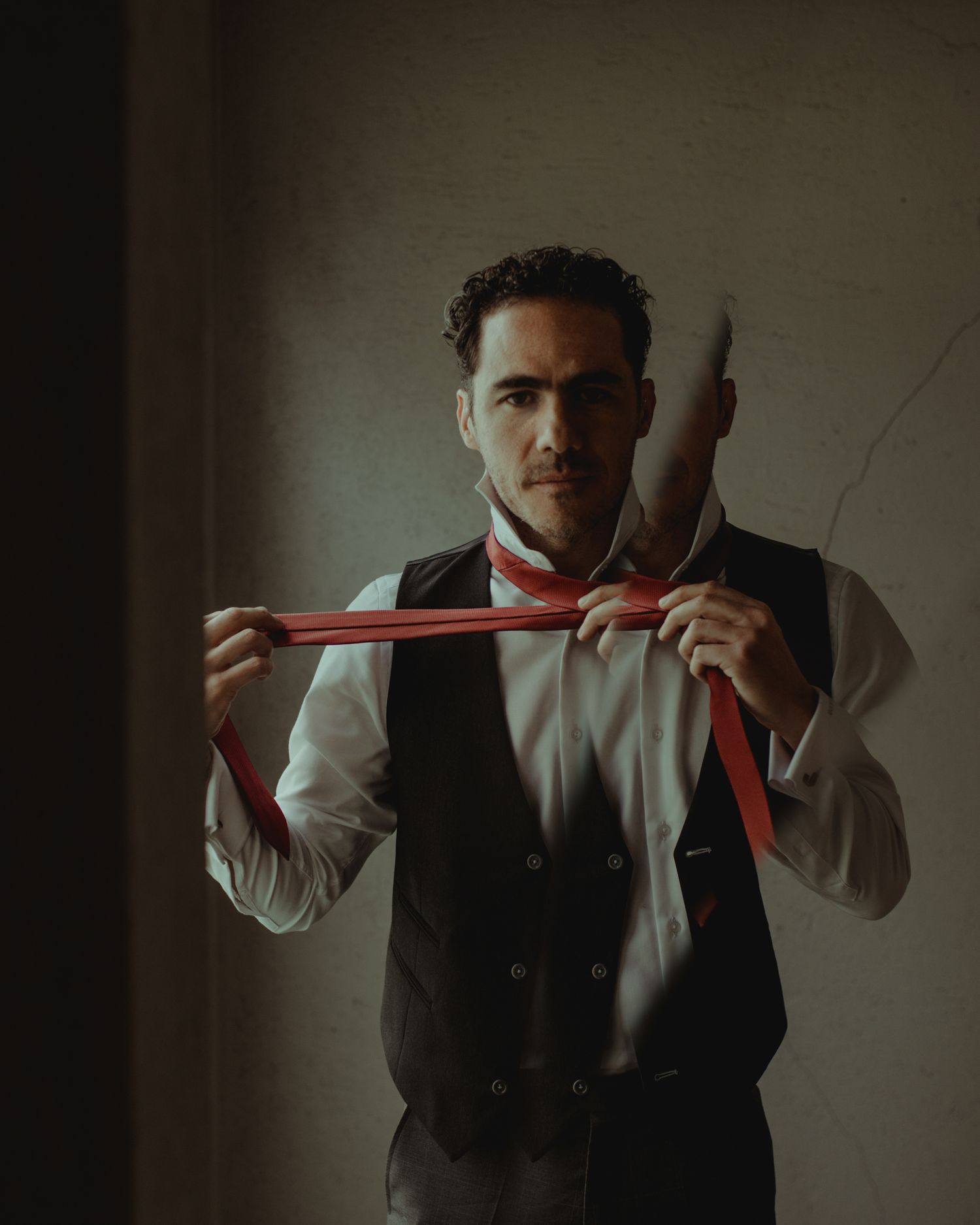 Person in formal attire holding red necktie while standing in moody lighting against wall.