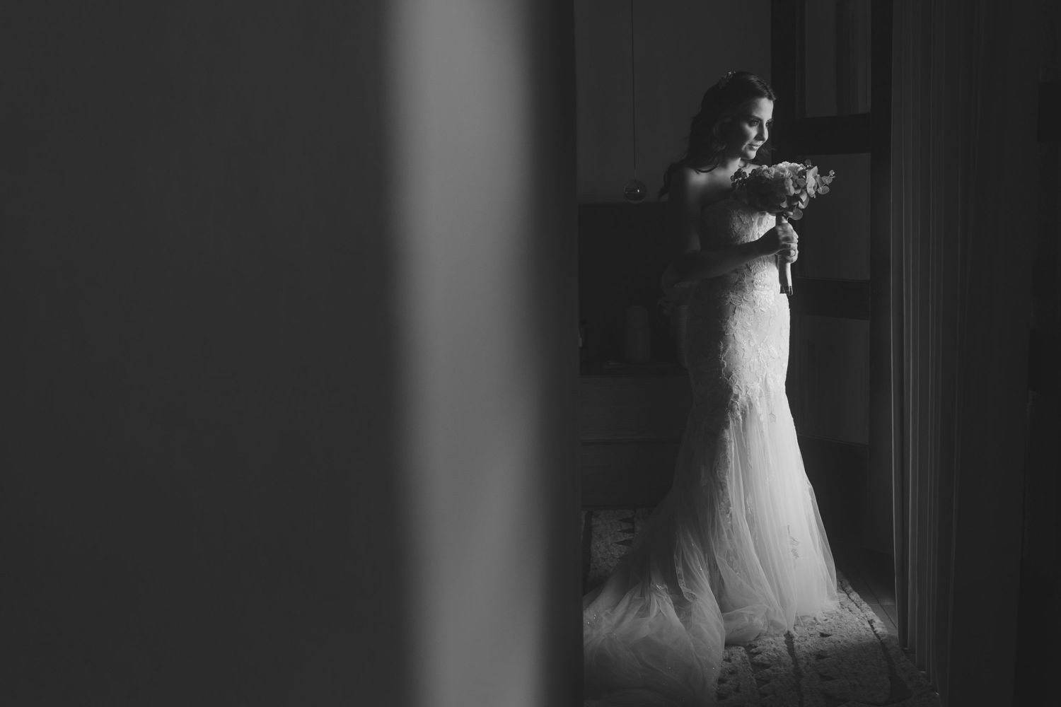 Black and white silhouette photo of a bride in a lace wedding gown standing by a window in dramatic lighting.