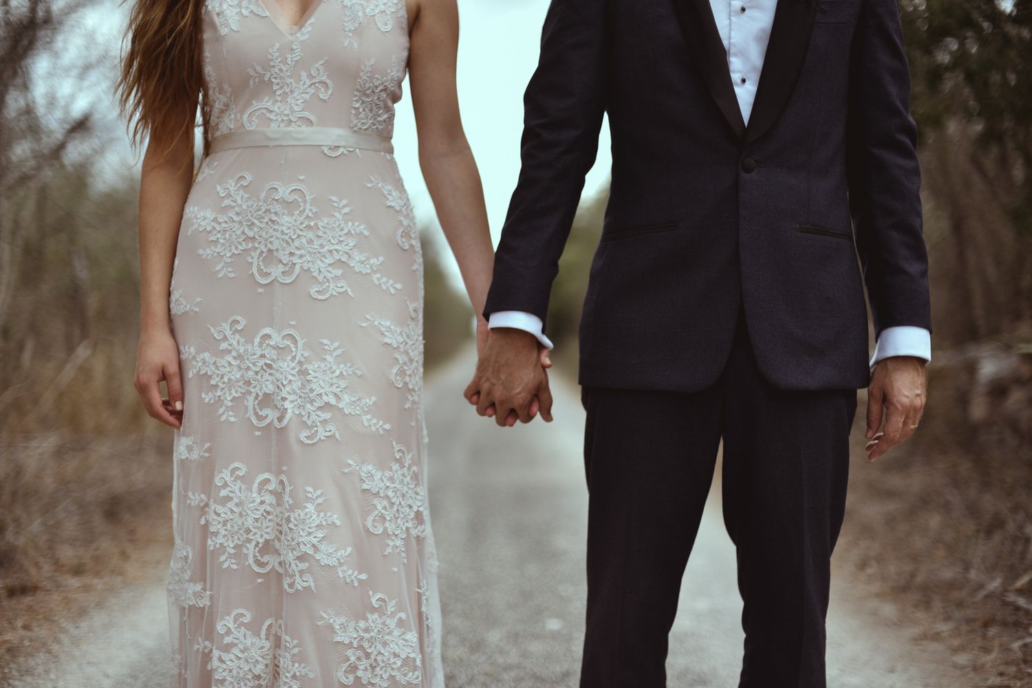 A couple in wedding attire holds hands on a woodland path, showing elegant lace gown and classic black suit details.