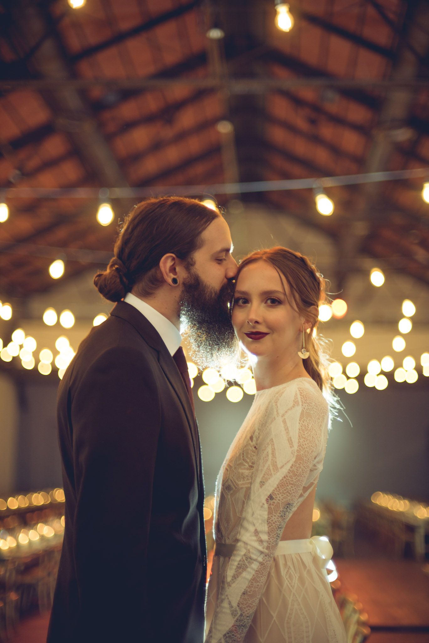 A romantic moment between newlyweds under warm string lights in a rustic barn venue with wooden beams overhead.