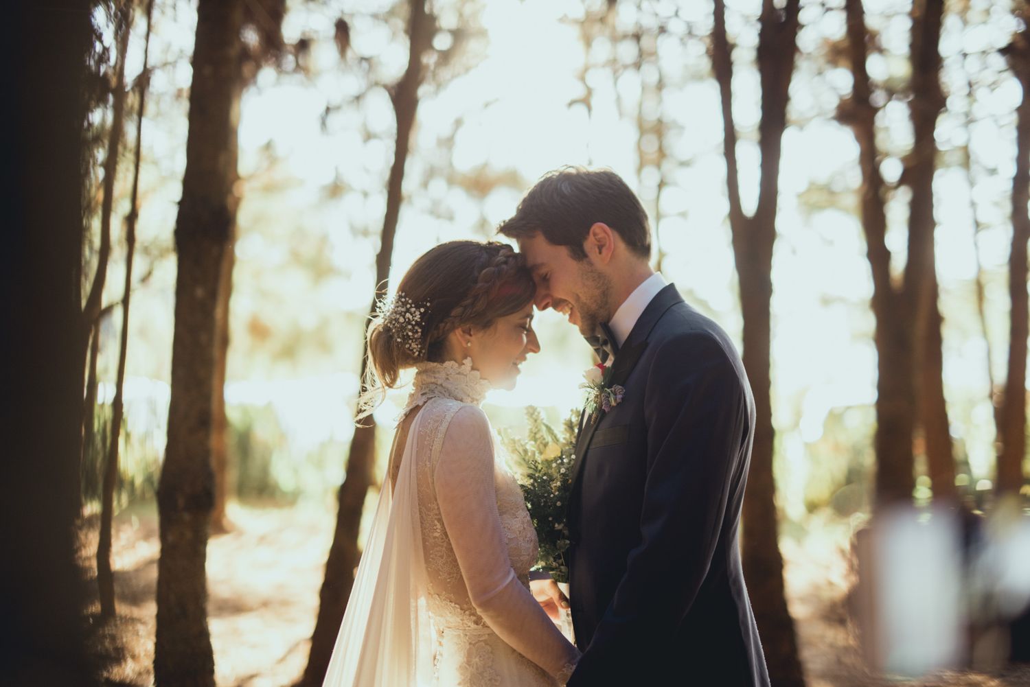 A romantic wedding couple shares an intimate moment in a sunlit forest during golden hour.