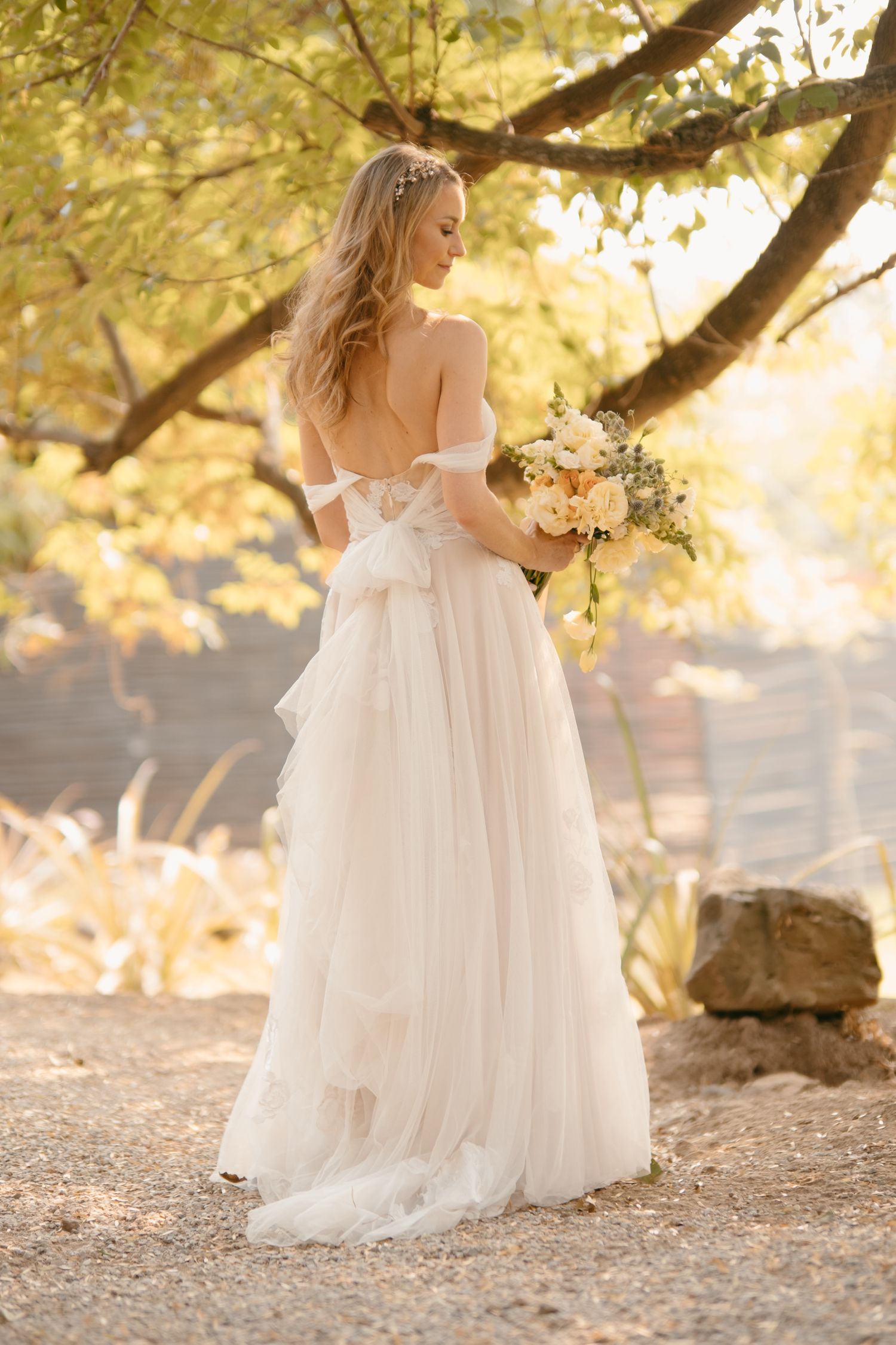 Bride in flowing white dress holds bouquet in golden sunset light beneath tree branches.