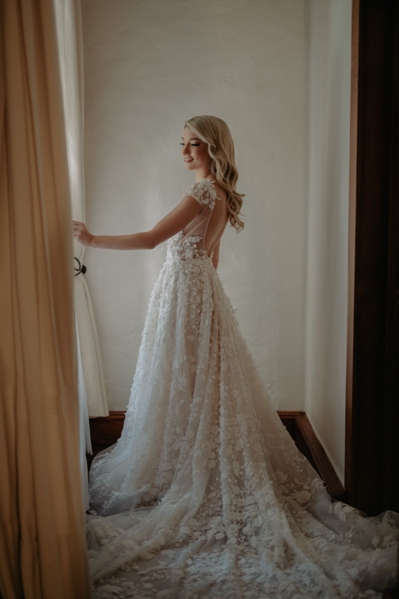 Bride in detailed lace ballgown poses by window in romantic bridal portrait.