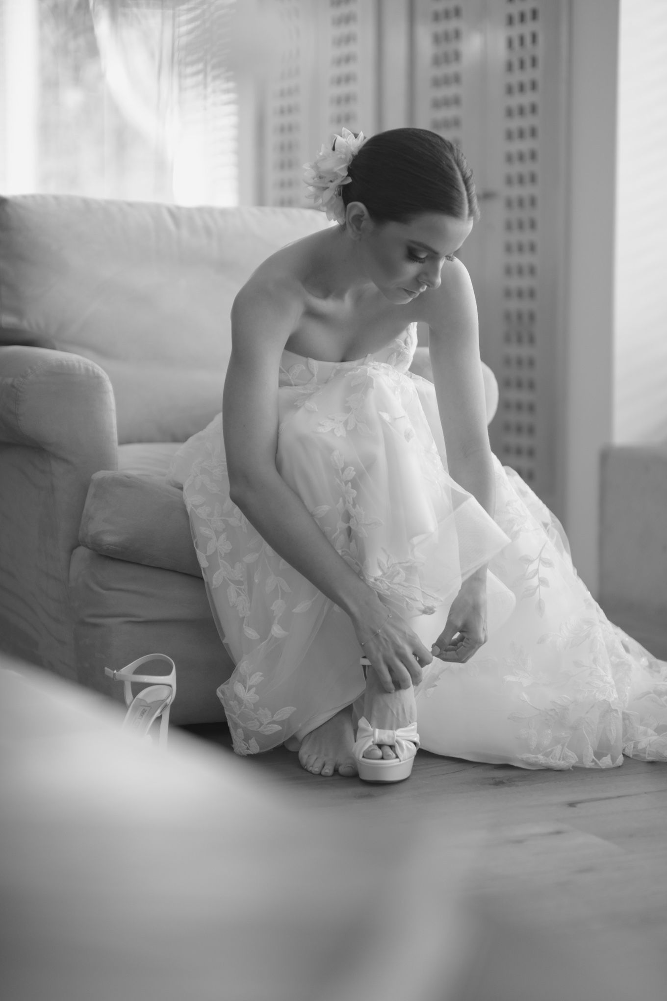A bride in a white wedding dress adjusts her shoe while sitting on a couch.