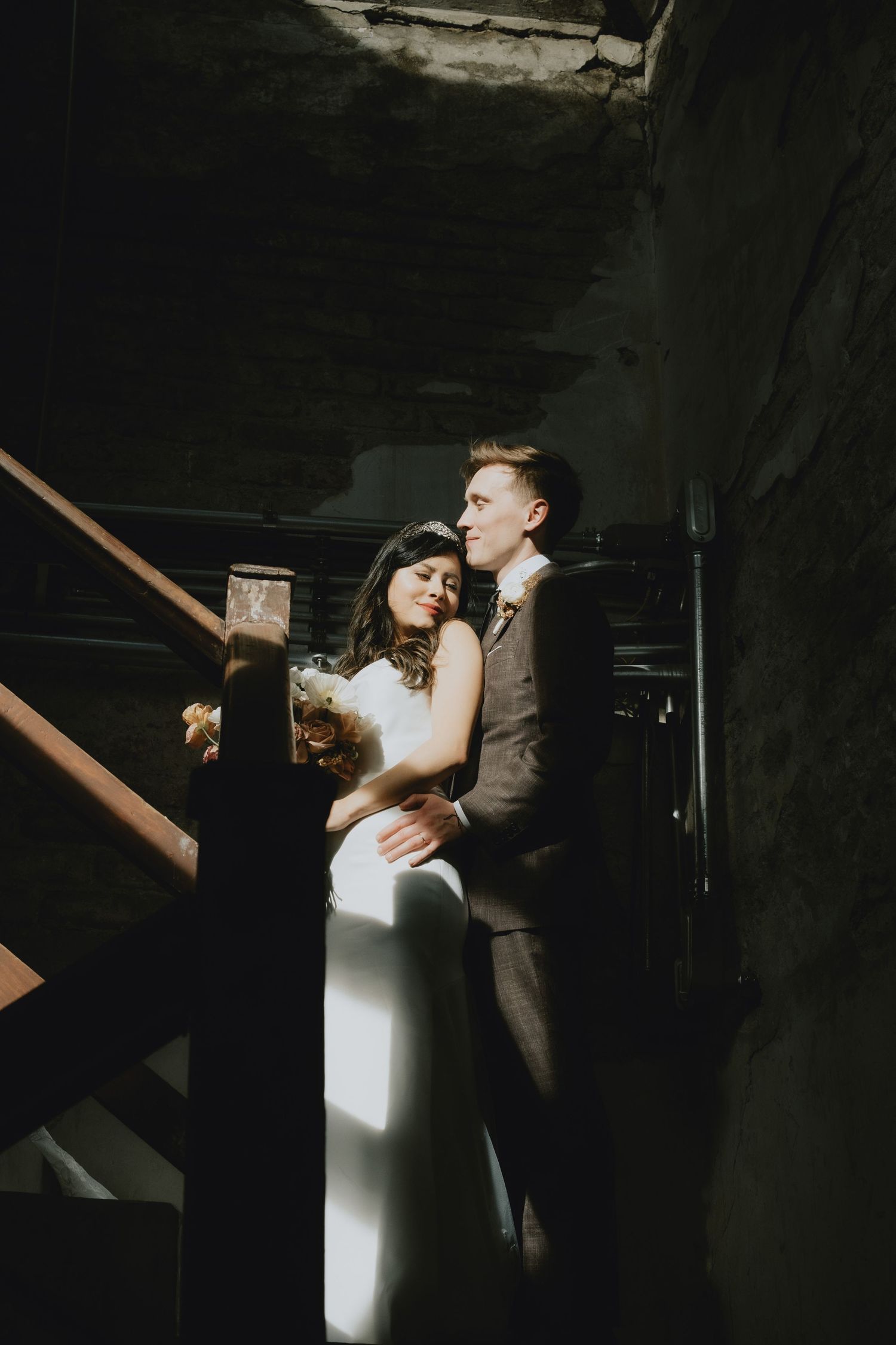 Dramatic wedding photo with couple in formal attire standing in dark stairwell with beam of natural light.
