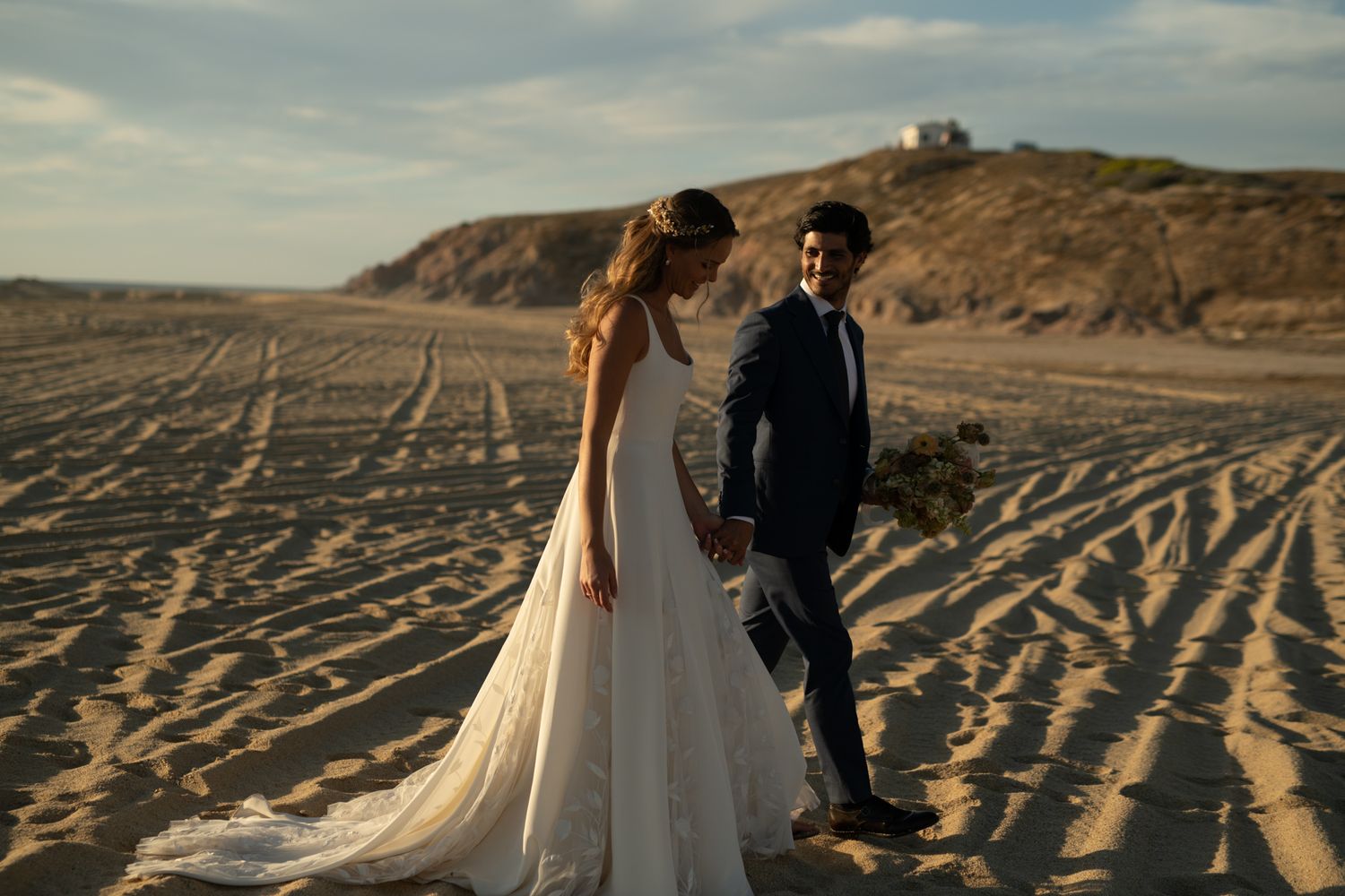 A wedding couple stands on sandy dunes during golden hour with hills in the background.