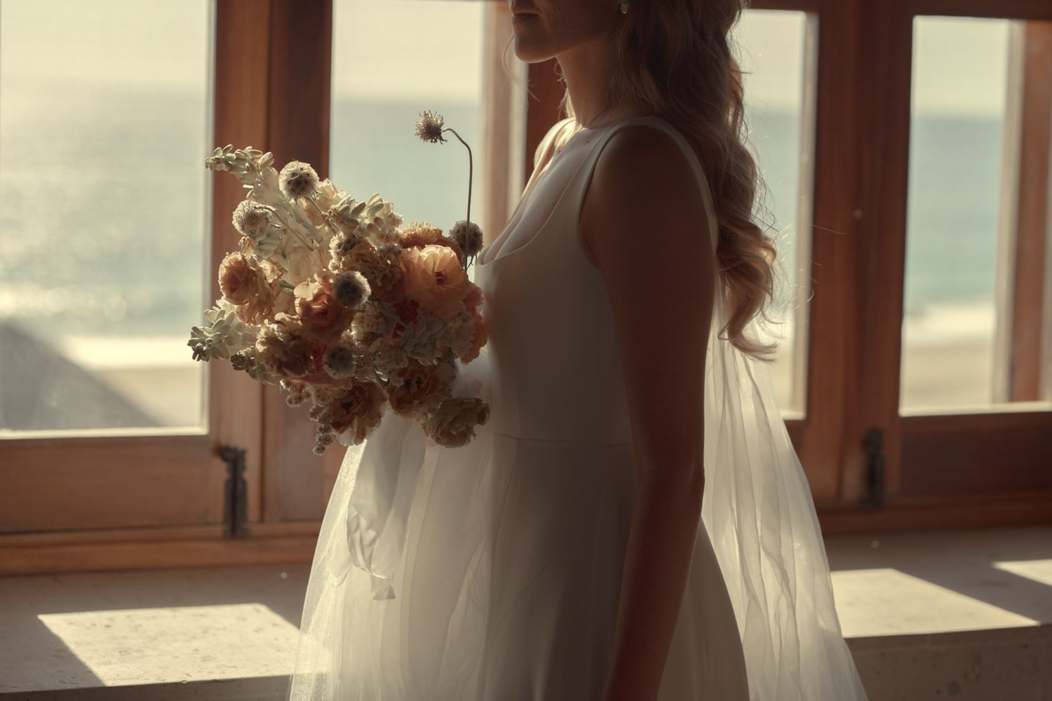 A bride in a white dress with long veil holds a pale bouquet while standing by sunlit windows in a dreamy setting.