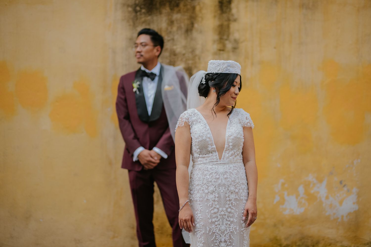 A couple in wedding attire stands against a textured yellow wall showing contrasting emotions.