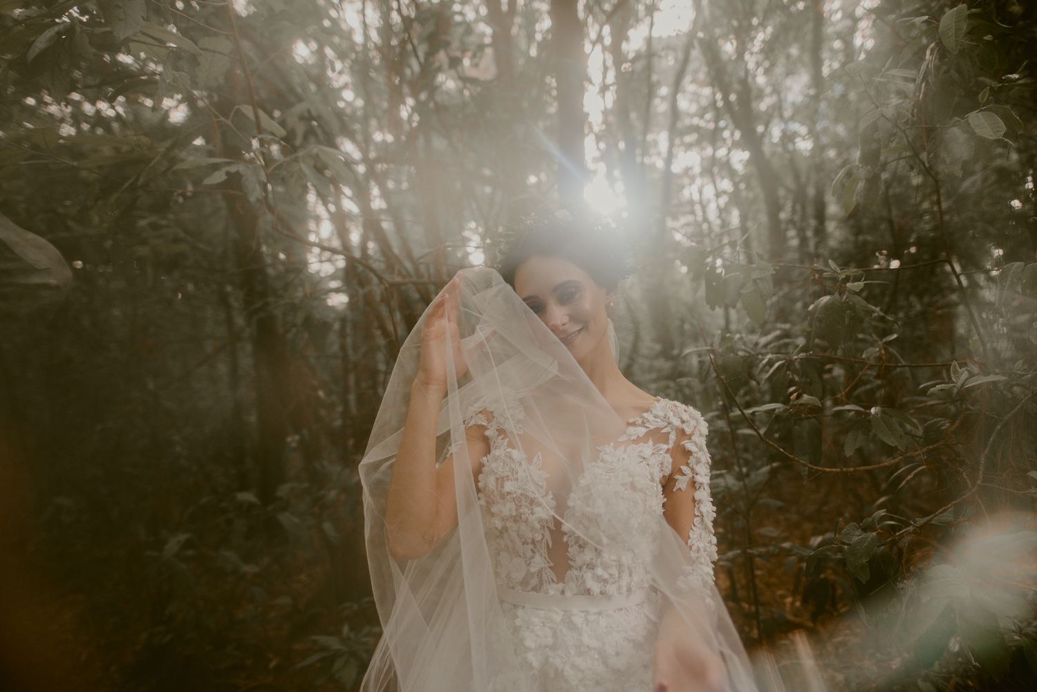 Sunlight streams through forest trees onto ethereal figure in delicate lace wedding gown with flowing veil.