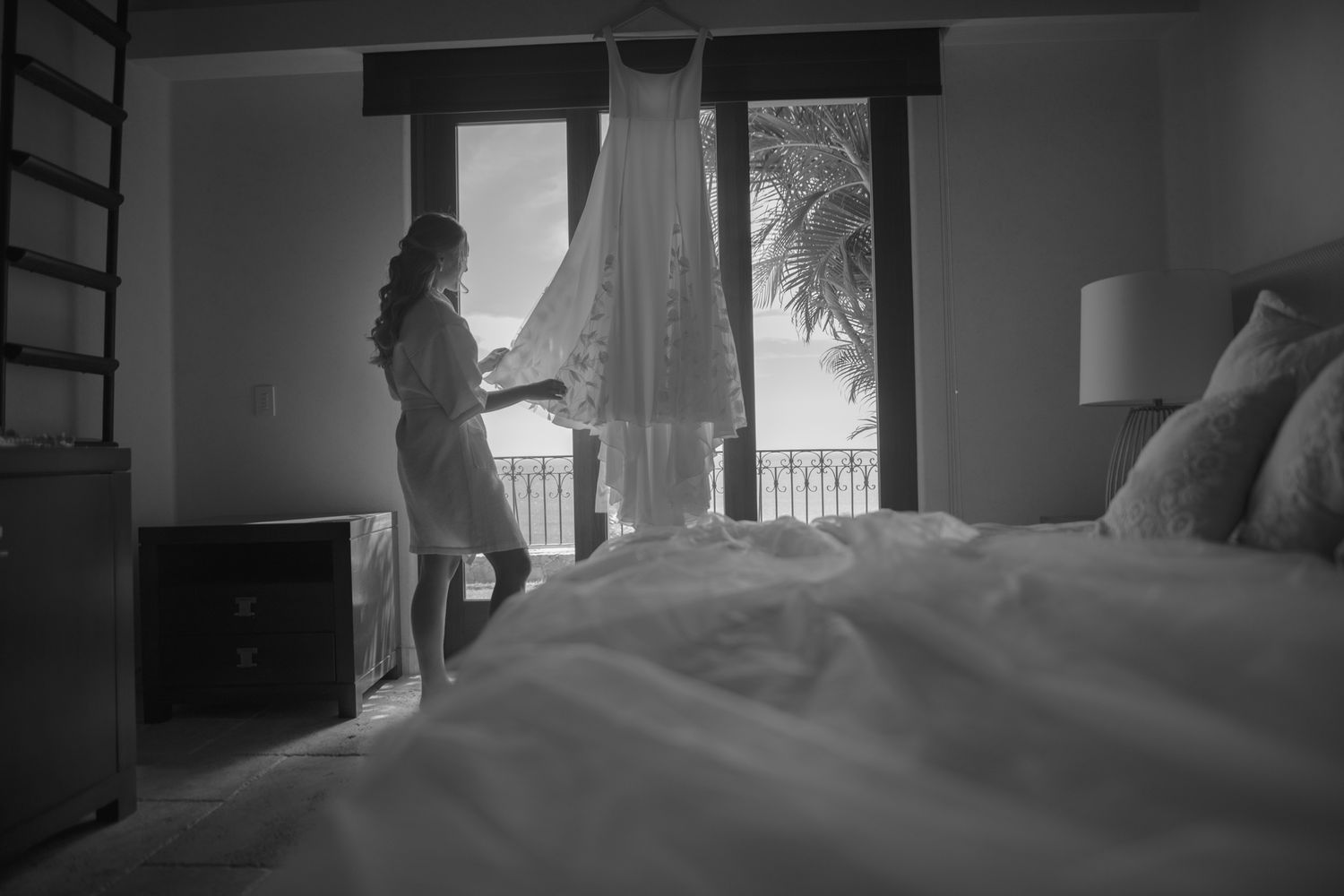 A silhouette stands by a window in a bright hotel room where a white dress hangs near a comfortable bed.