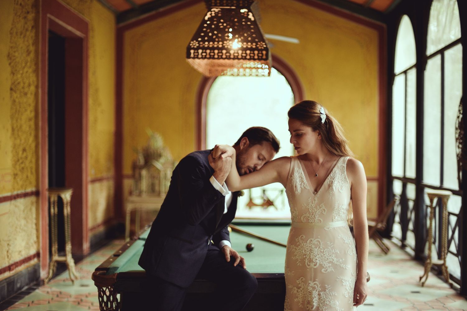 A romantic couple in wedding attire share an intimate moment in an elegant hallway with yellow walls and ornate lighting.