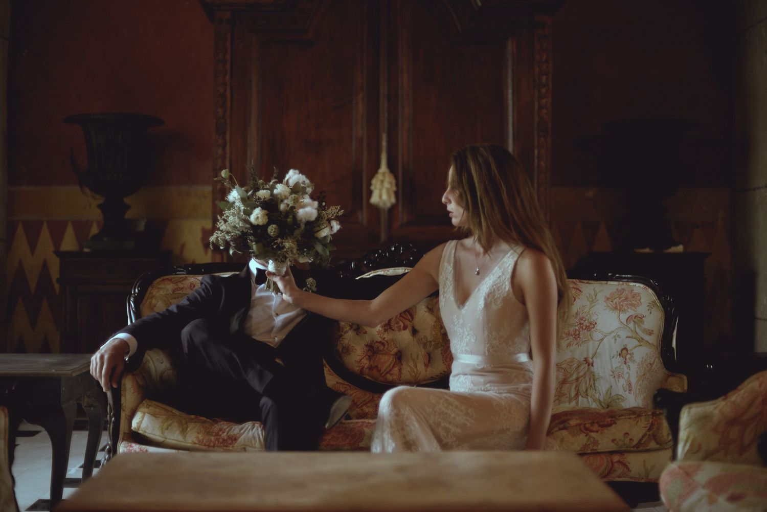 Two people sit in a dimly lit room on a vintage sofa with floral upholstery next to a vase of white flowers.