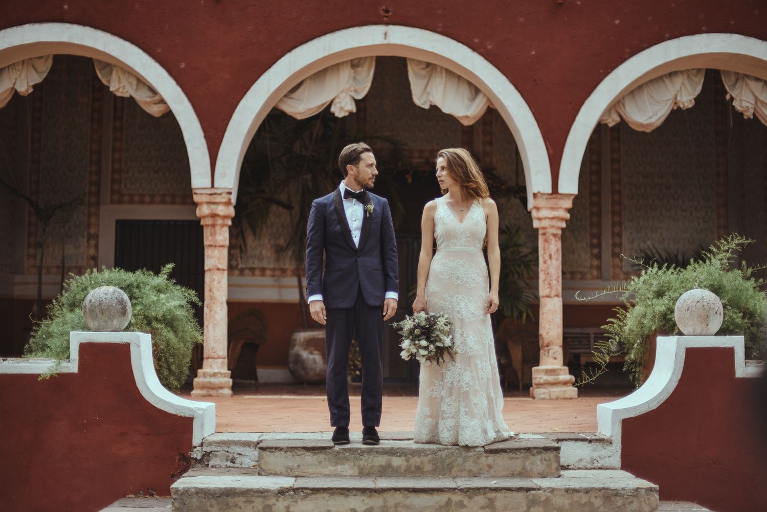 A couple stands on stone steps beneath elegant arched columns and coral-colored walls at an upscale hacienda venue.