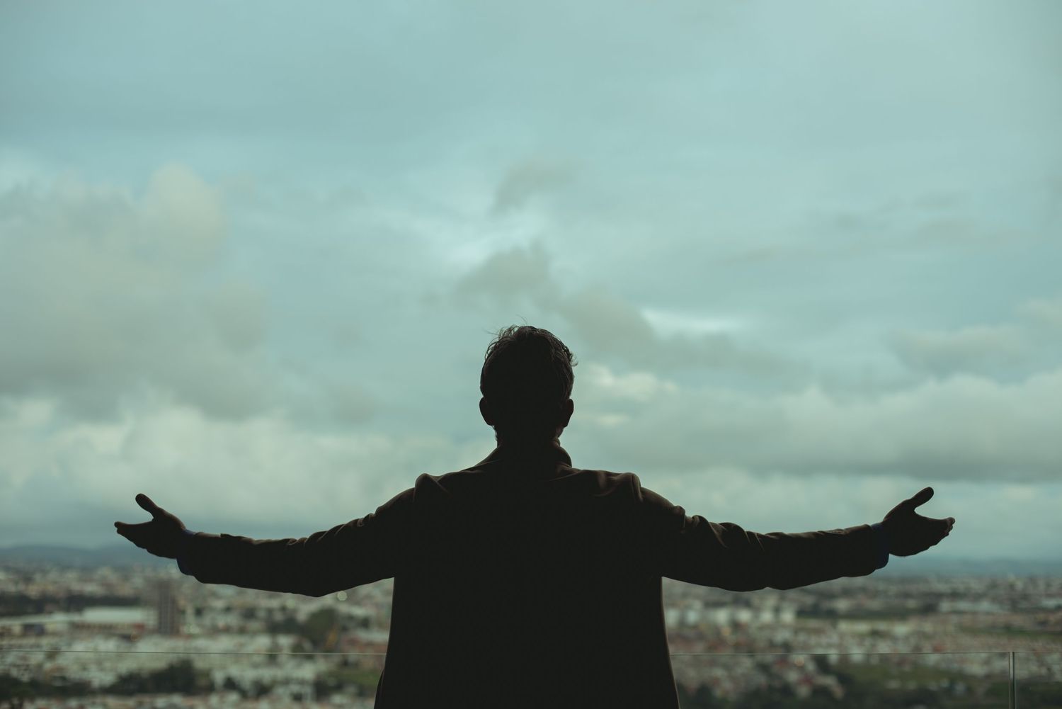 Silhouette of person with arms spread wide against cloudy cityscape.