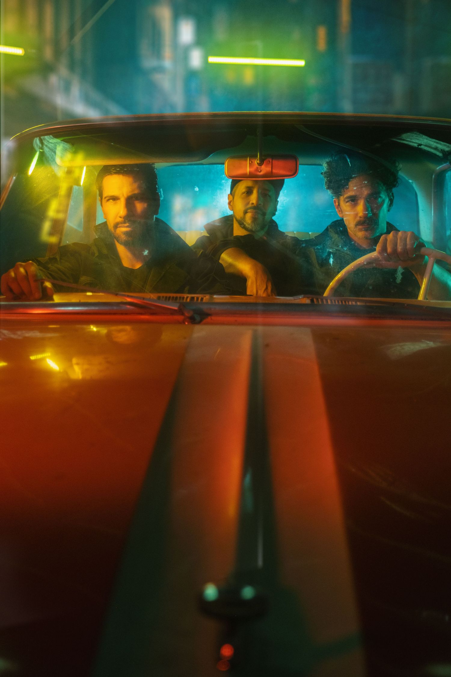 Three people sit in a car at night illuminated by neon lights casting vibrant blue and yellow glows through the windows.